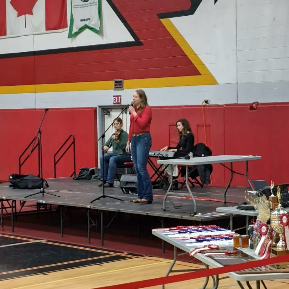 A female speaker standing on a stage with a microphone at an indoor event, with two women seated behind her, one playing keyboard; background features red and white walls with flags and awards on a table nearby.
