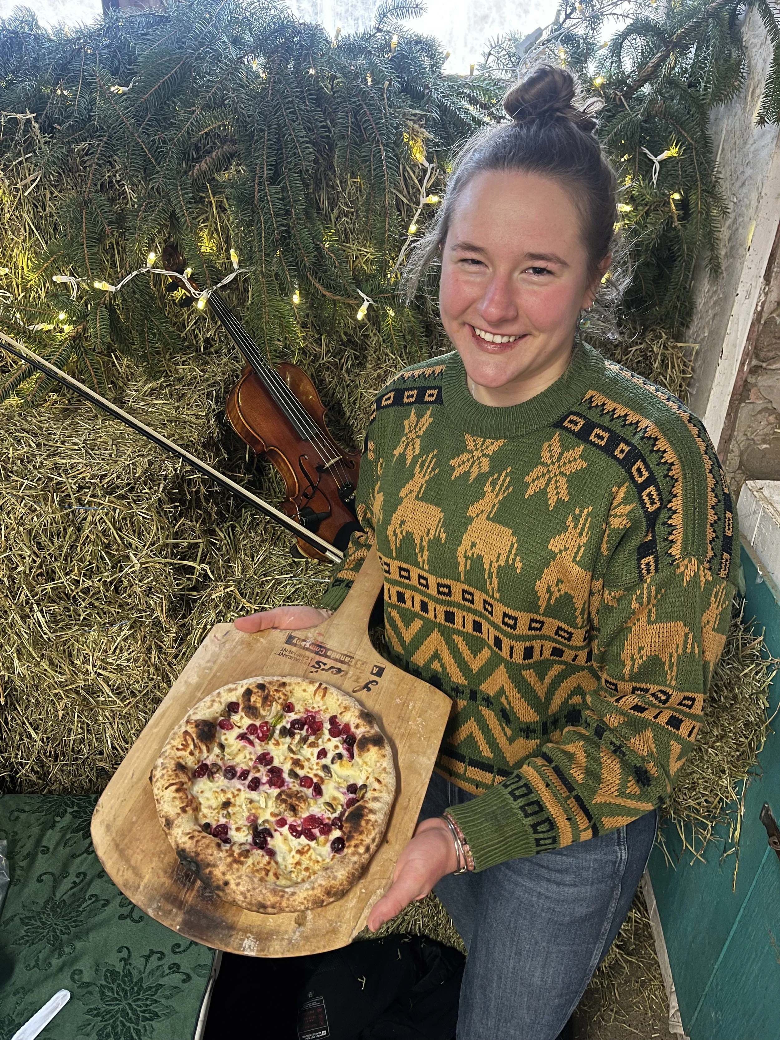 A smiling woman with a top bun hairstyle holding a wooden pizza peel with a freshly baked pizza topped with cheese and pomegranate seeds. In the background, there are evergreen branches, string lights, and a violin leaning against the foliage.