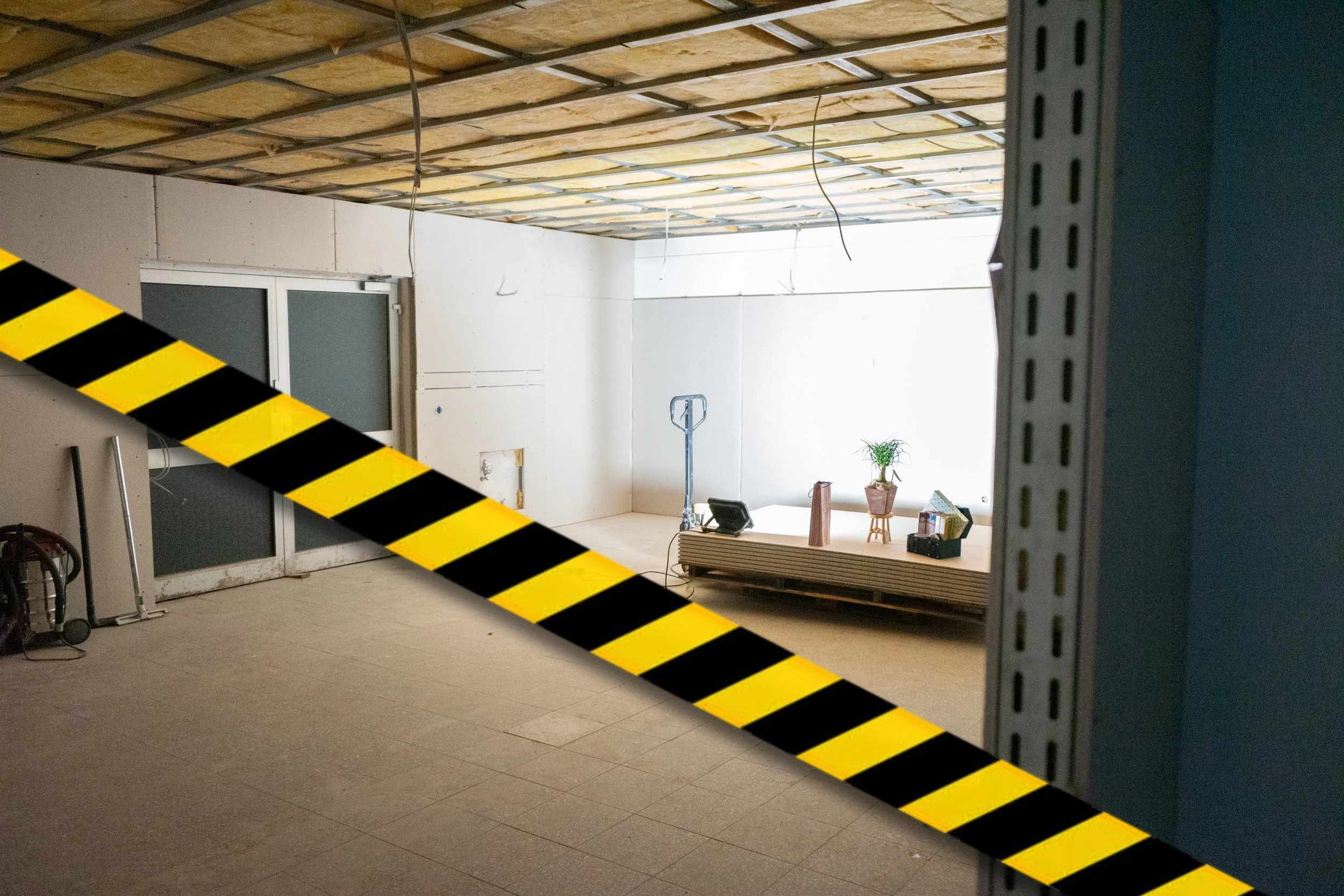 Innenraum einer Bauhalle mit Bauarbeiten im Gange, im Hintergrund sind Bauwerkzeuge wie eine Säge, eine Wasserwaage und Bauplatten, sowie eine Pflanze auf einer Kiste zu sehen, im Vordergrund eine gelb-schwarze Absperrung.