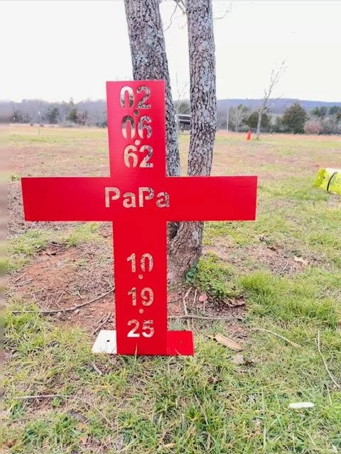 A red cross-shaped memorial marker with dates and the word "Papa" in the center, placed outdoors on grass with trees and open field in the background.