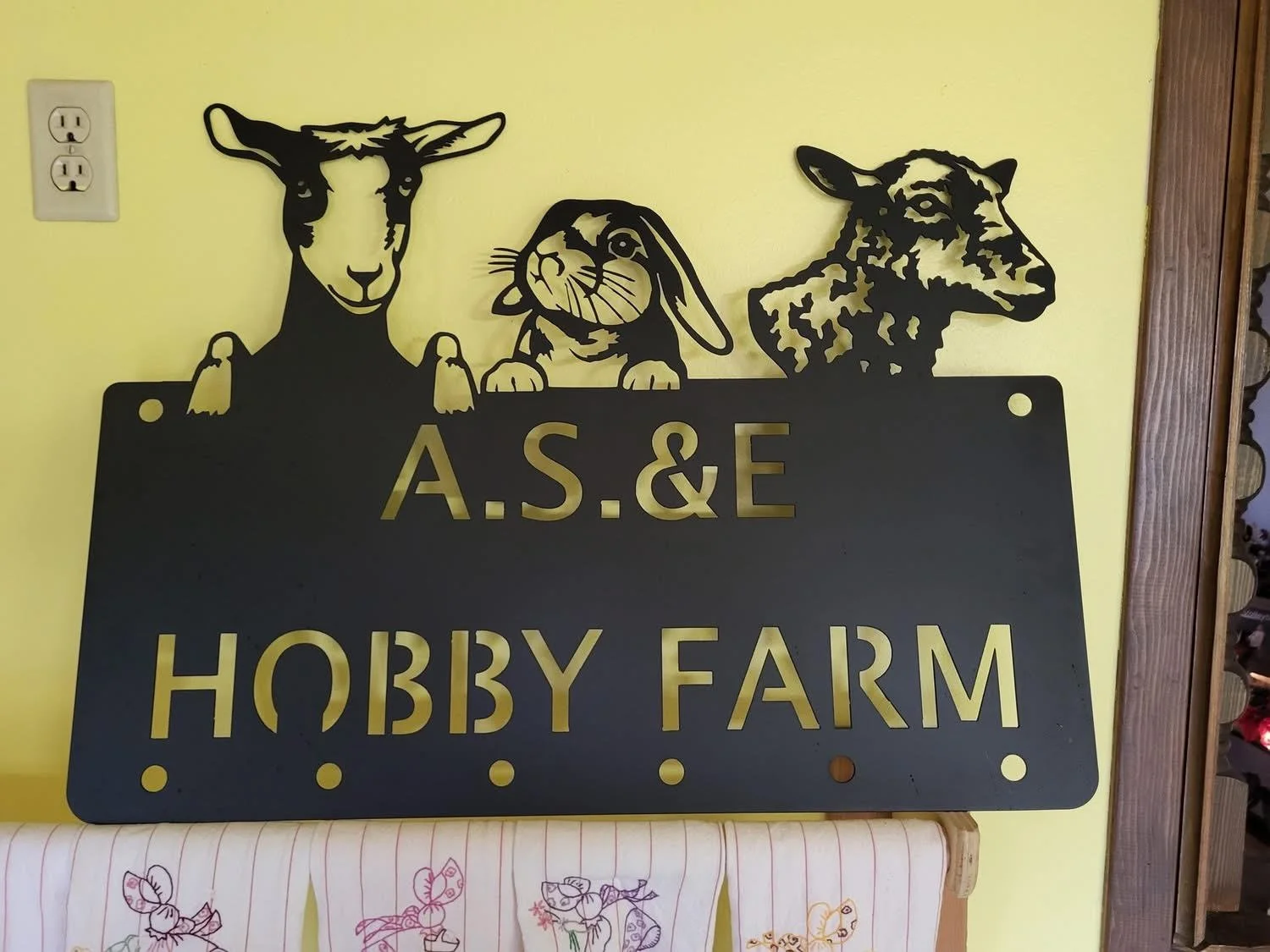 Black metal sign with three animal silhouettes and yellow lettering that reads "A.S. & E Hobby Farm". The animals are a goat on the left, a rabbit in the middle, and a dog on the right, flying on top of the sign.