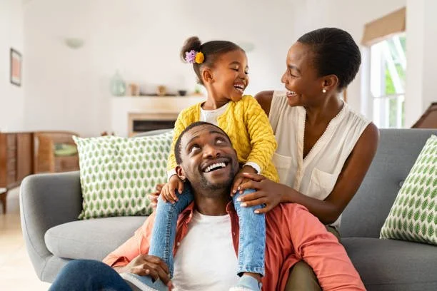 A family of three, a man, woman, and young girl, smiling and enjoying time together on a gray couch in a bright living room.