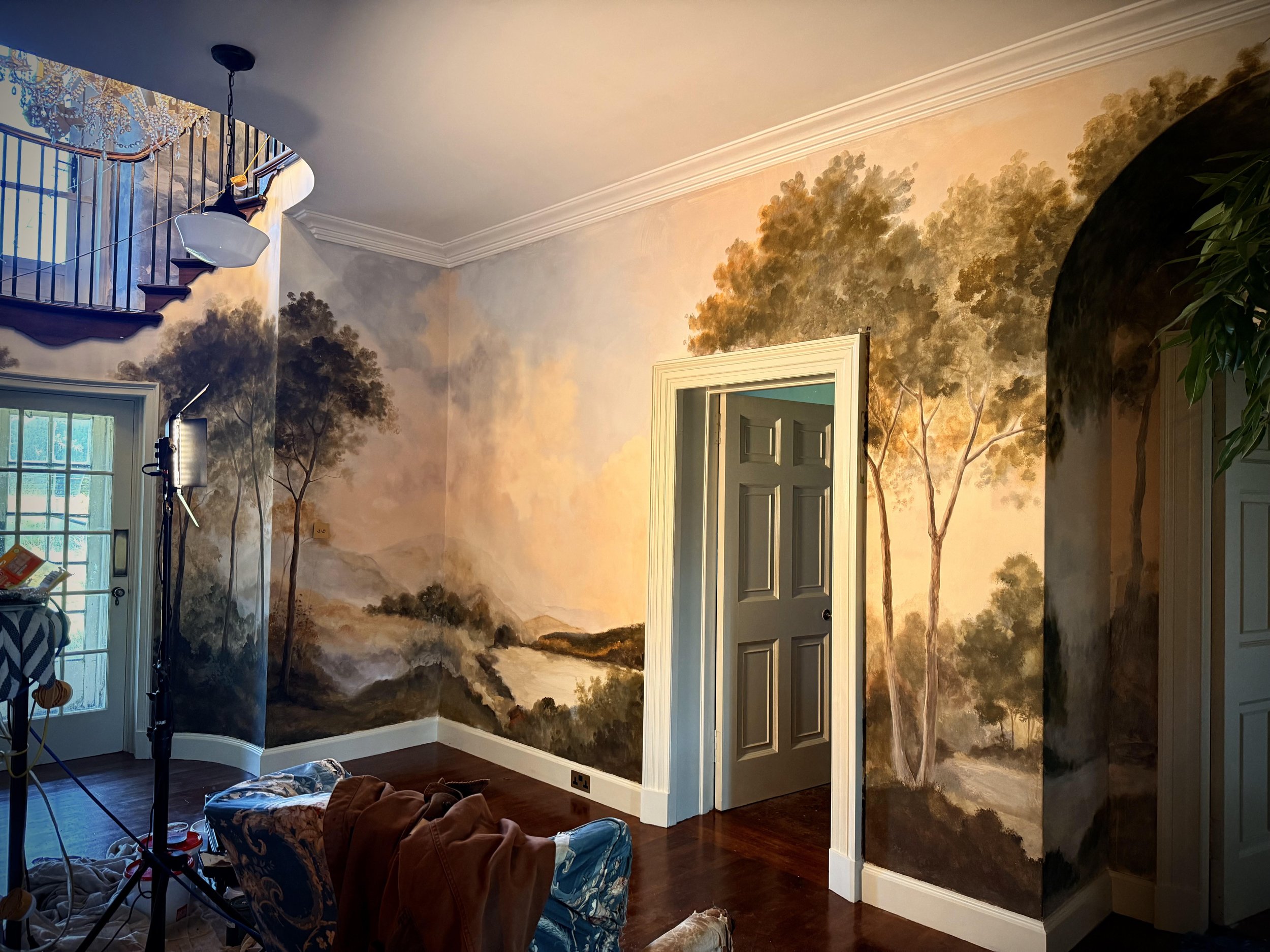 Interior of a room with a large landscape mural of trees and a river, a closed door, hardwood floors, and a sliding glass door leading outside.