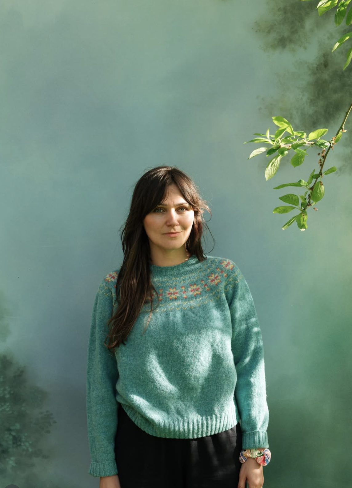 A woman with long dark hair and light skin wearing a green sweater with a floral pattern on the chest, standing outdoors in front of a green wall with some foliage in the upper right corner.