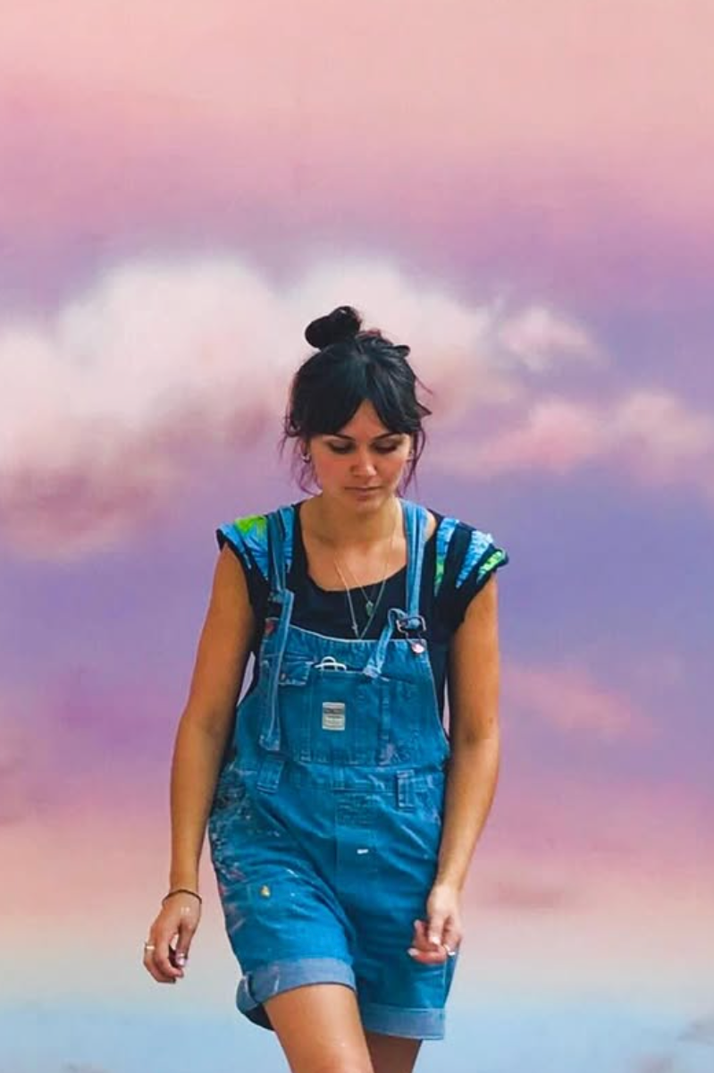 Young woman with dark hair tied in a bun, wearing denim overalls and a black t-shirt, walking outdoors against a colorful sky with pink and purple clouds.