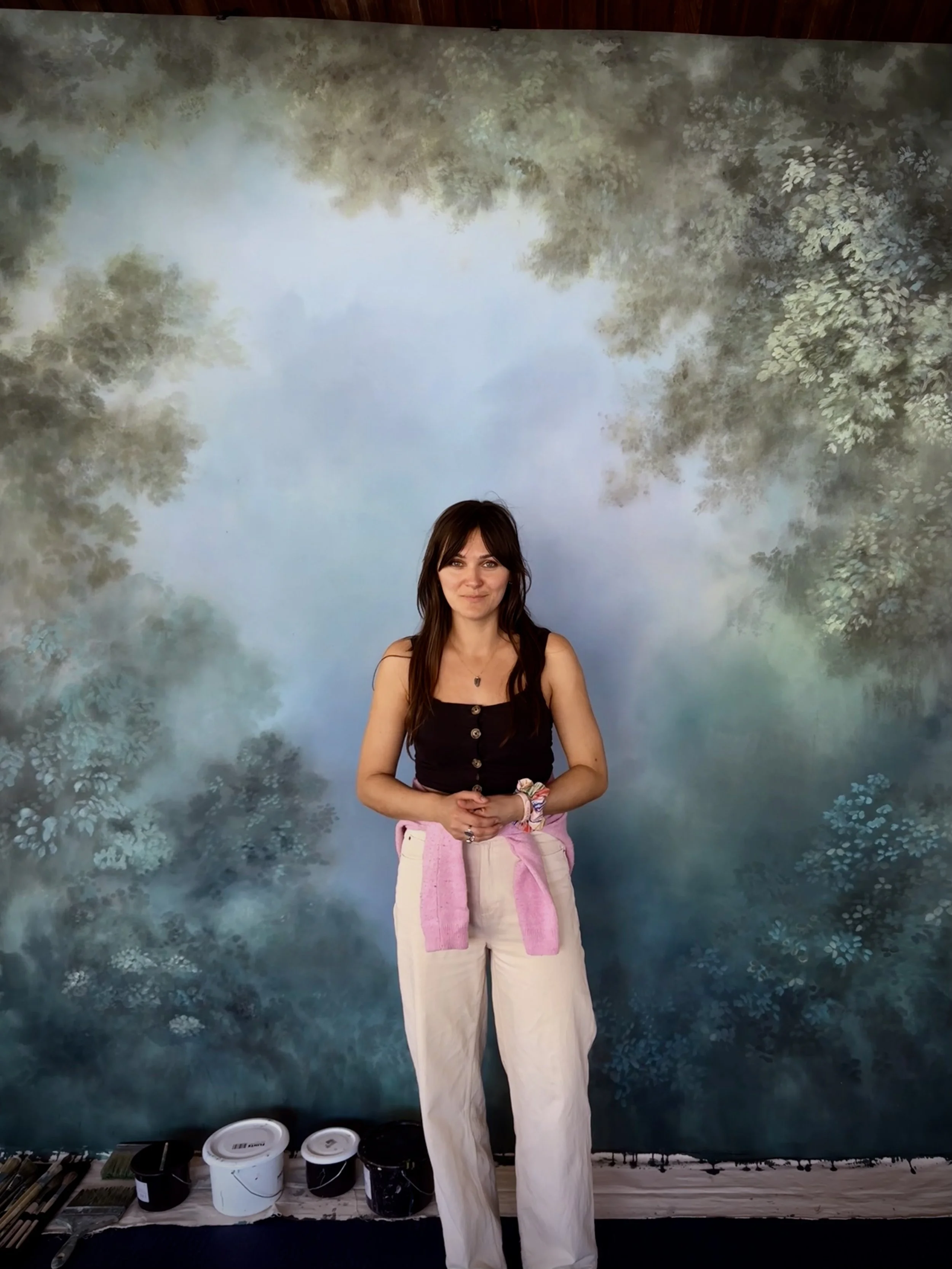 A woman with long dark hair, wearing a black sleeveless top and beige pants, stands in front of a painted backdrop of a forest scene with trees and sky. She is smiling slightly and has a pink sweater tied around her waist. There are paint buckets and brushes on the floor in front of her.