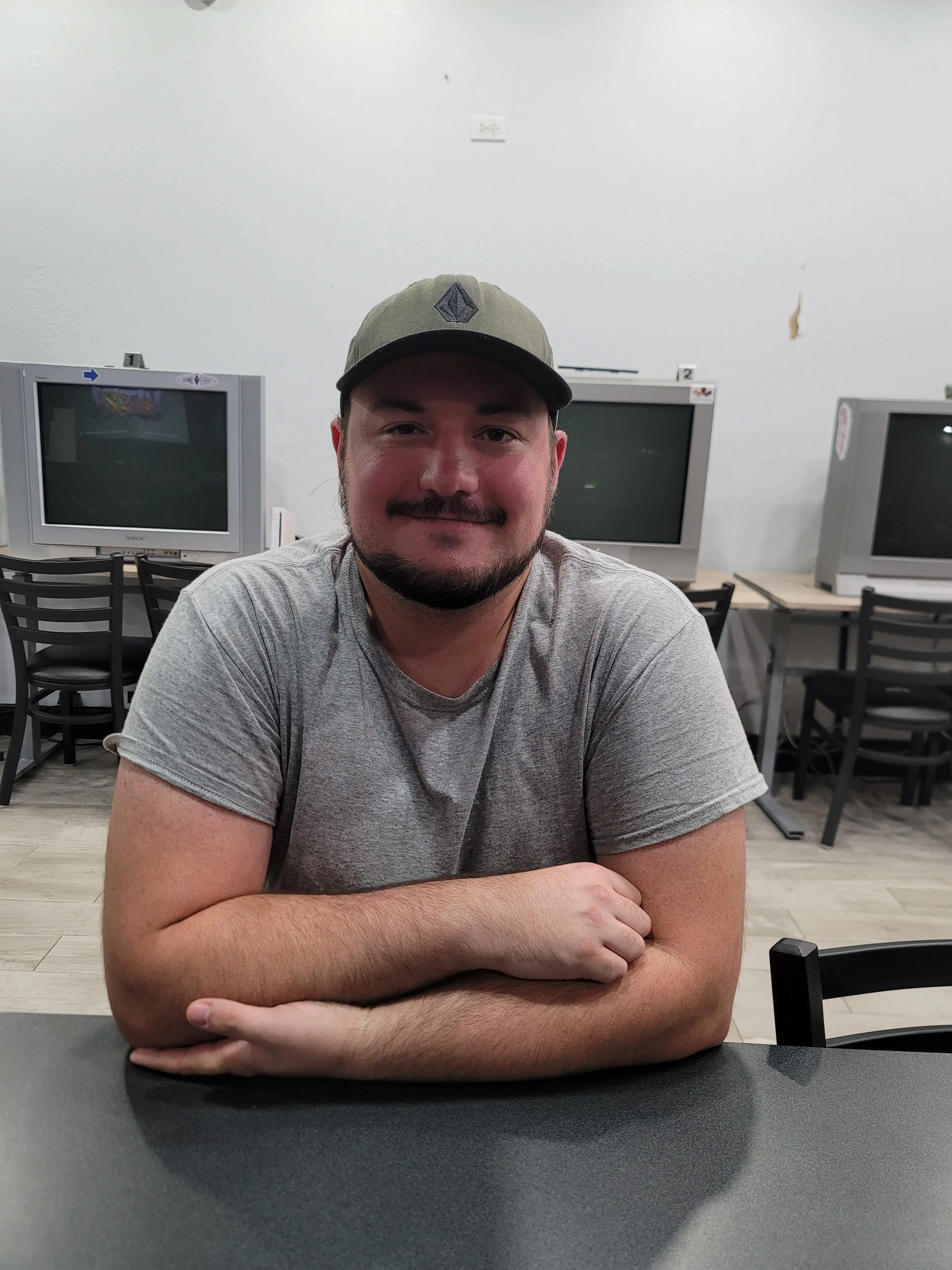 A young man with a beard and mustache wearing a gray T-shirt and a green baseball cap sitting at a table in a room with three old televisions in the background.