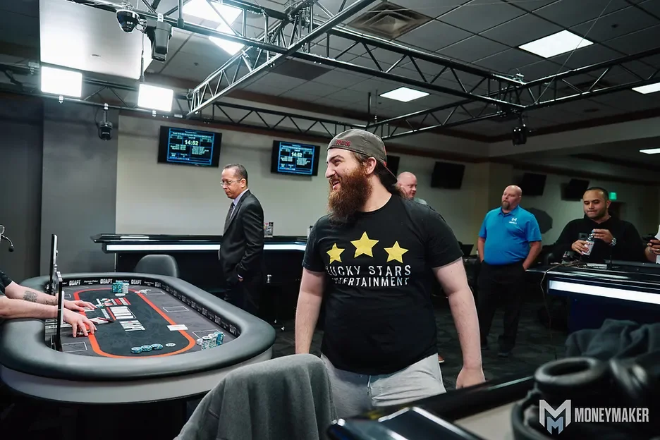 A man with a beard and wearing a black T-shirt with gold stars and the text 'Rocky Stars Entertainment' stands at a poker table in a casino, smiling and looking towards another person who is sitting at the table. Other people are watching, some taking photos, in a well-lit room with digital screens on the wall, a casino style poker table, and the Moneymaker logo visible.