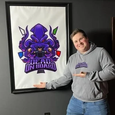 A young man in a gray hoodie points to a large framed poster with a purple octopus and the text 'Dead on Board' on the wall.