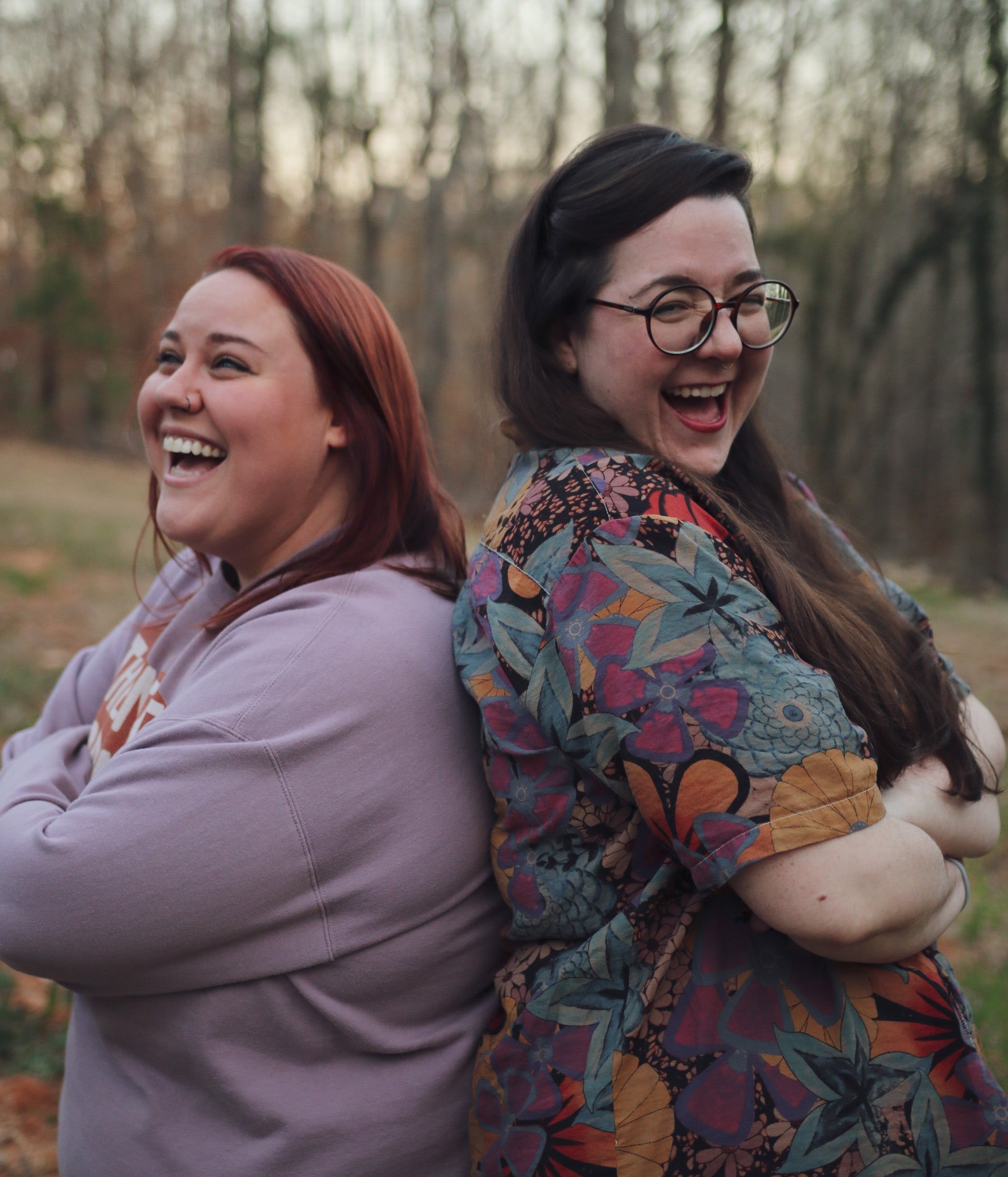 Two women standing back to back outdoors, smiling and laughing in a wooded area.