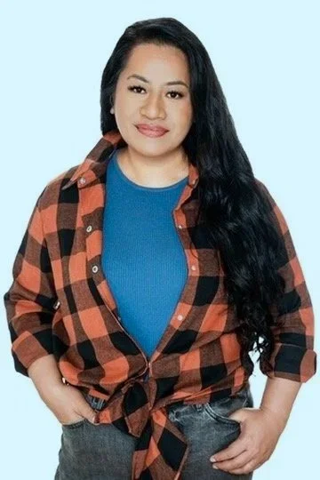 Young woman with long black hair, wearing a blue shirt and an orange and black checkered flannel tied at the waist, standing against a light blue background.