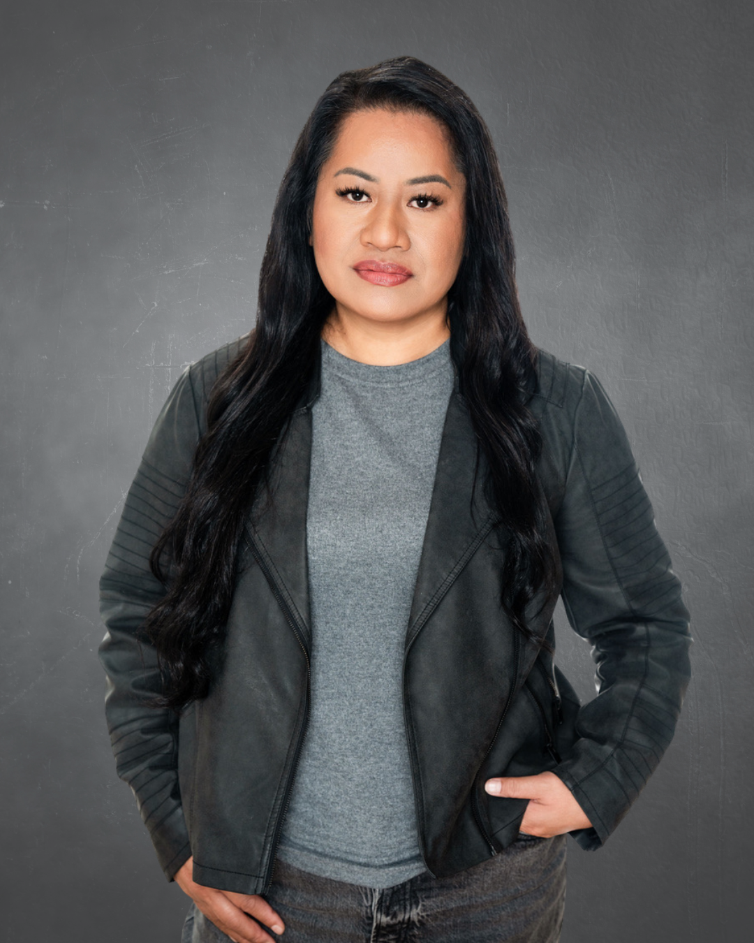A woman with long black hair wearing a gray T-shirt and black leather jacket stands against a gray textured background.