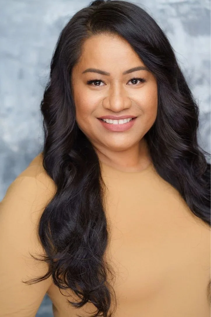 A woman with long, wavy black hair, wearing a beige top, smiling, with a blurred gray background.