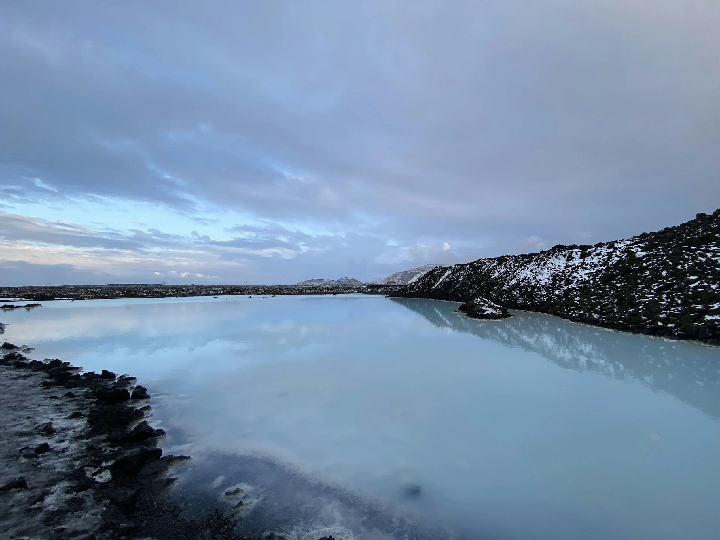A calm body of water reflecting a cloudy blue sky, surrounded by snow-covered hills and dark rocky shores.