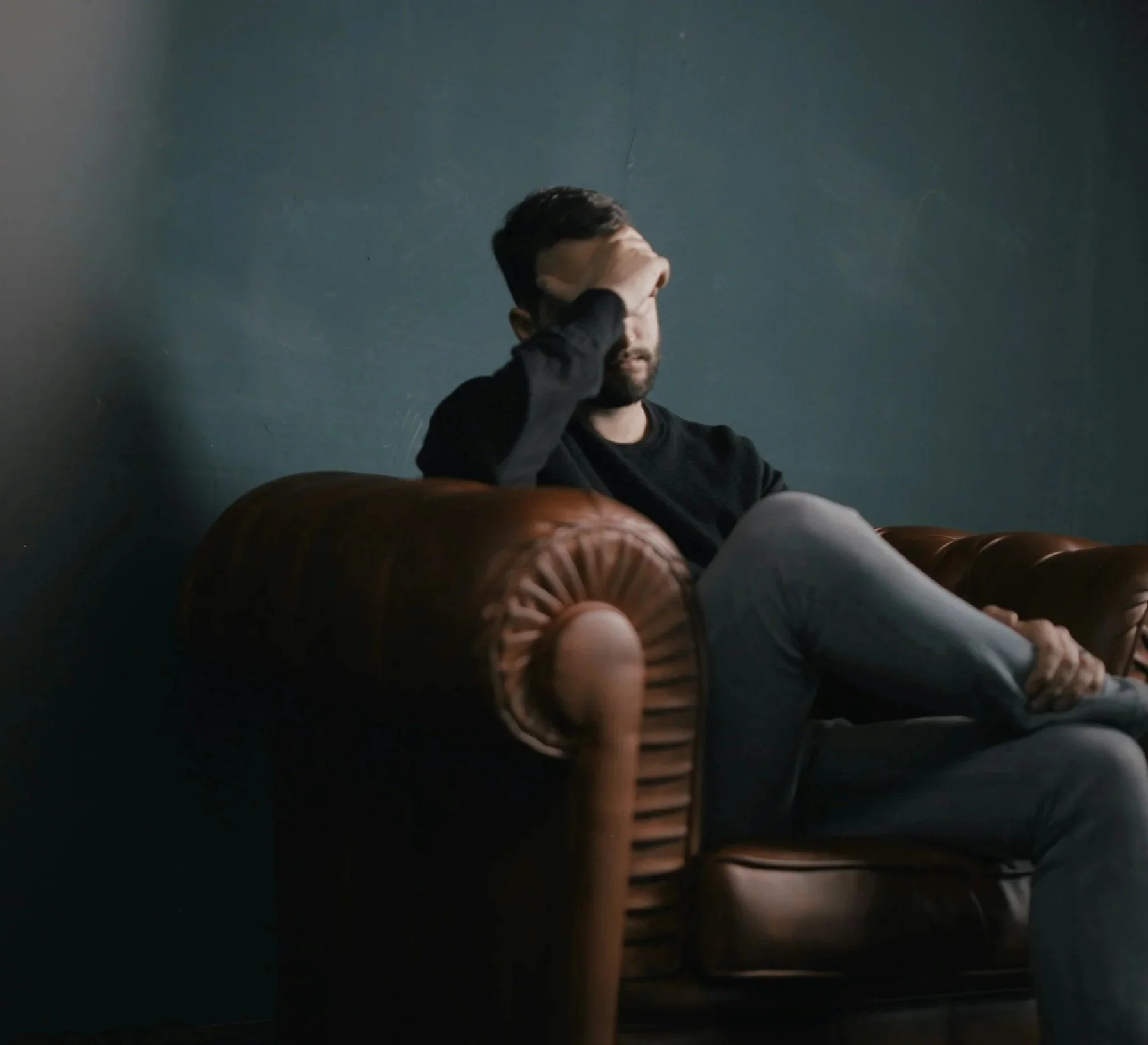 A man sitting on a brown leather couch against a dark blue wall, holding his head with his right hand, appearing thoughtful or distressed.
