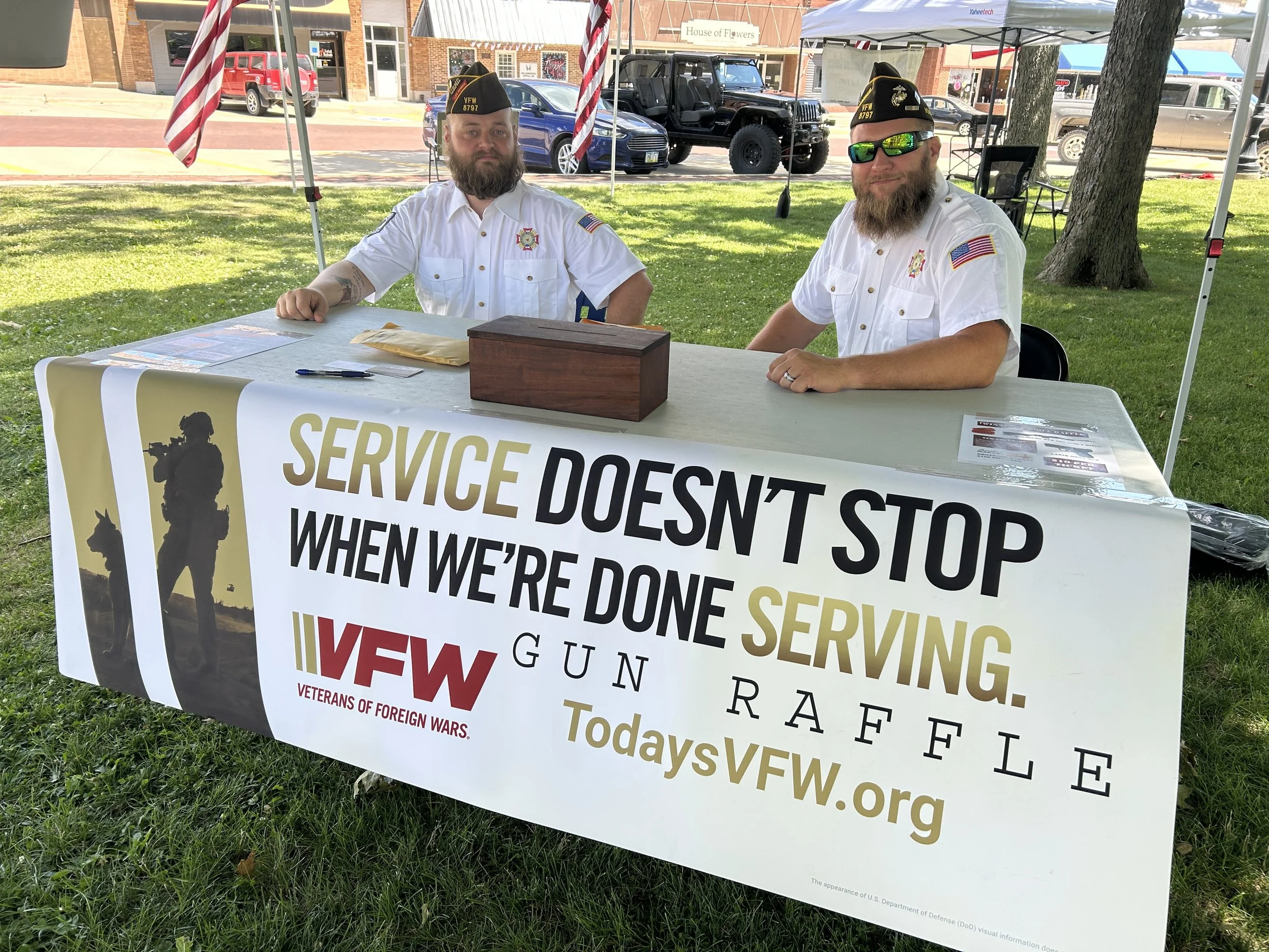 Two men in white shirts and black hats sitting at a table under a park canopy, with a large sign promoting veterans and service members, reading 'Service doesn't stop when we're done serving. GUN RAFE TodayVFW.org,' and an American flag in the background.