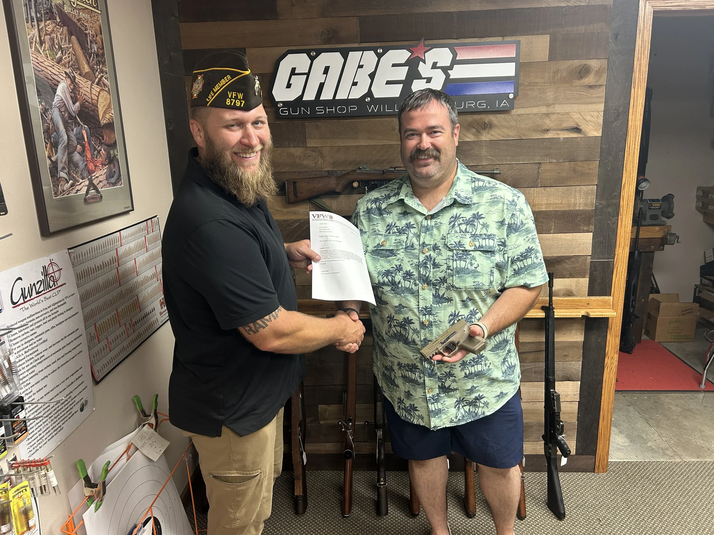 Two men shake hands inside a gun shop, one holding a handgun and a document. There are rifles in the background, with a sign reading 'GABE'S GUN SHOP' on the wall.