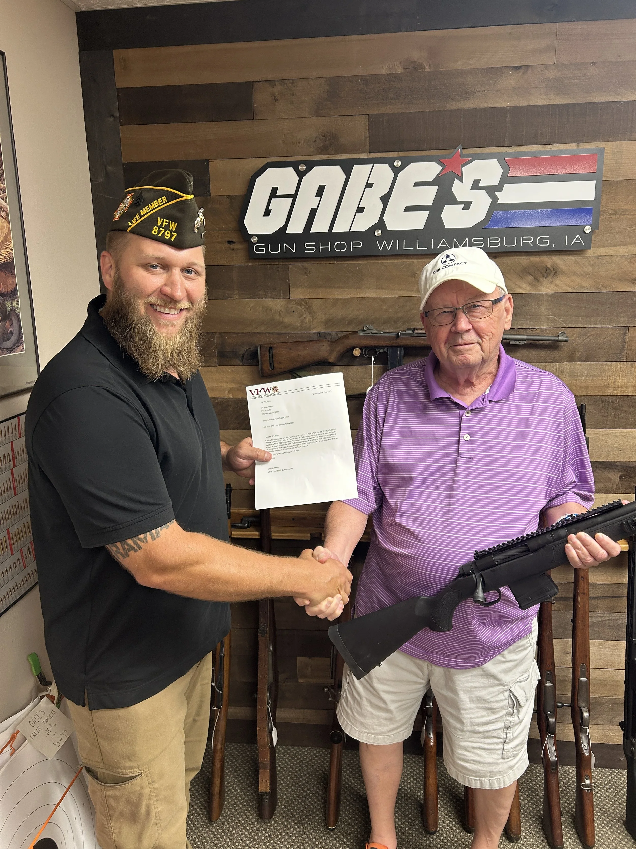 Two men shaking hands inside a gun shop, one young man with a beard wearing a VFW cap and a black shirt, the other older man in a white cap and purple striped shirt holding a rifle. They are in front of a wooden wall with a sign that says "GABE'S GUN SHOP WILLIAMSBURG, IA."
