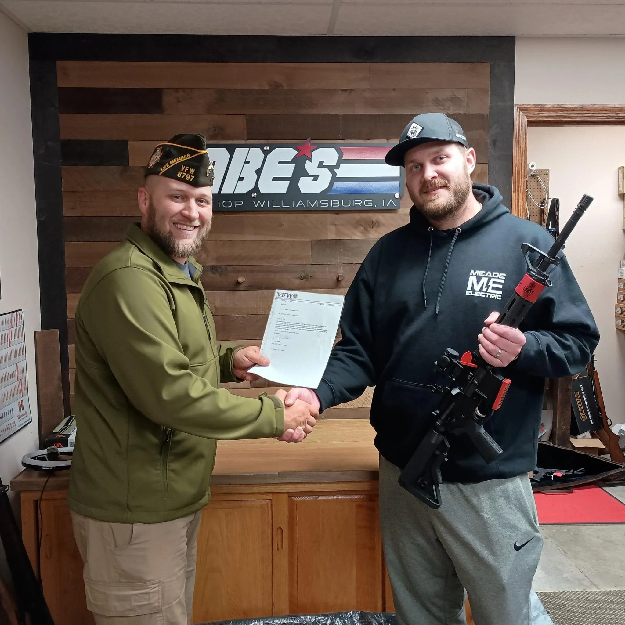 Two men shake hands inside a shop, with one man holding a document and the other holding a rifle. They are smiling. The shop's sign behind them reads 'VETS SHOP WILLIAMSBURG, IA.'