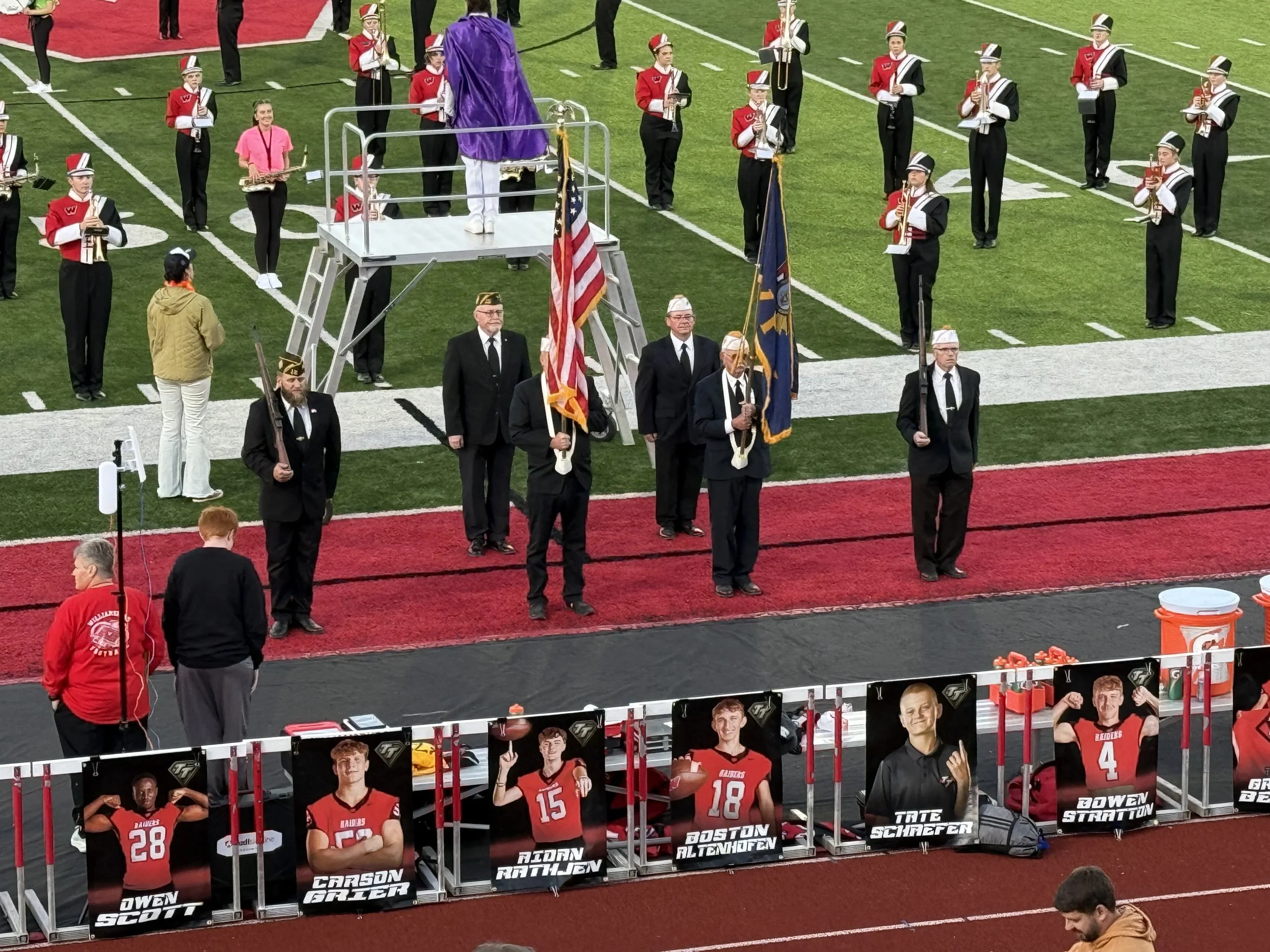 A football field with a marching band, individuals holding flags, and people standing behind numbered posters of football players.