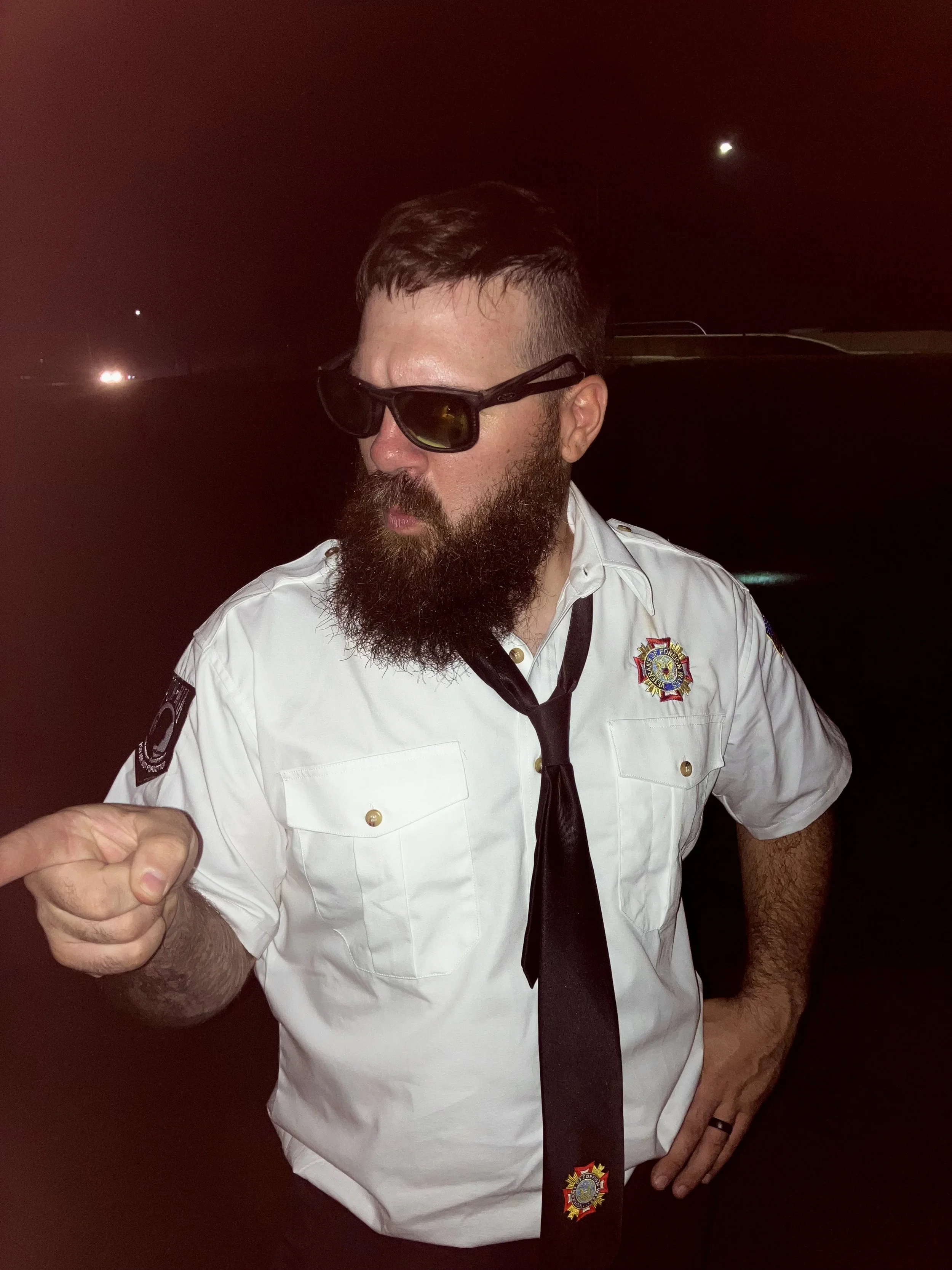 A man dressed as a firefighter, wearing sunglasses, a white firefighter uniform with badge, black tie, and a ring on his finger, standing at night.