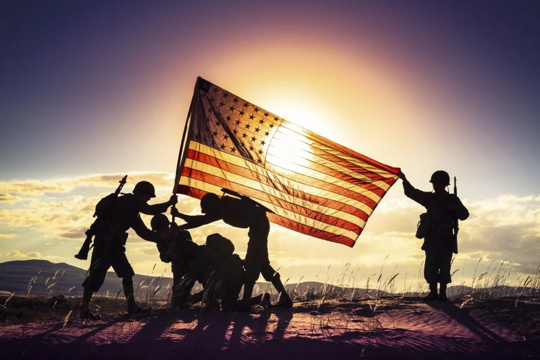 Silhouettes of soldiers raising the American flag at sunset in a field.