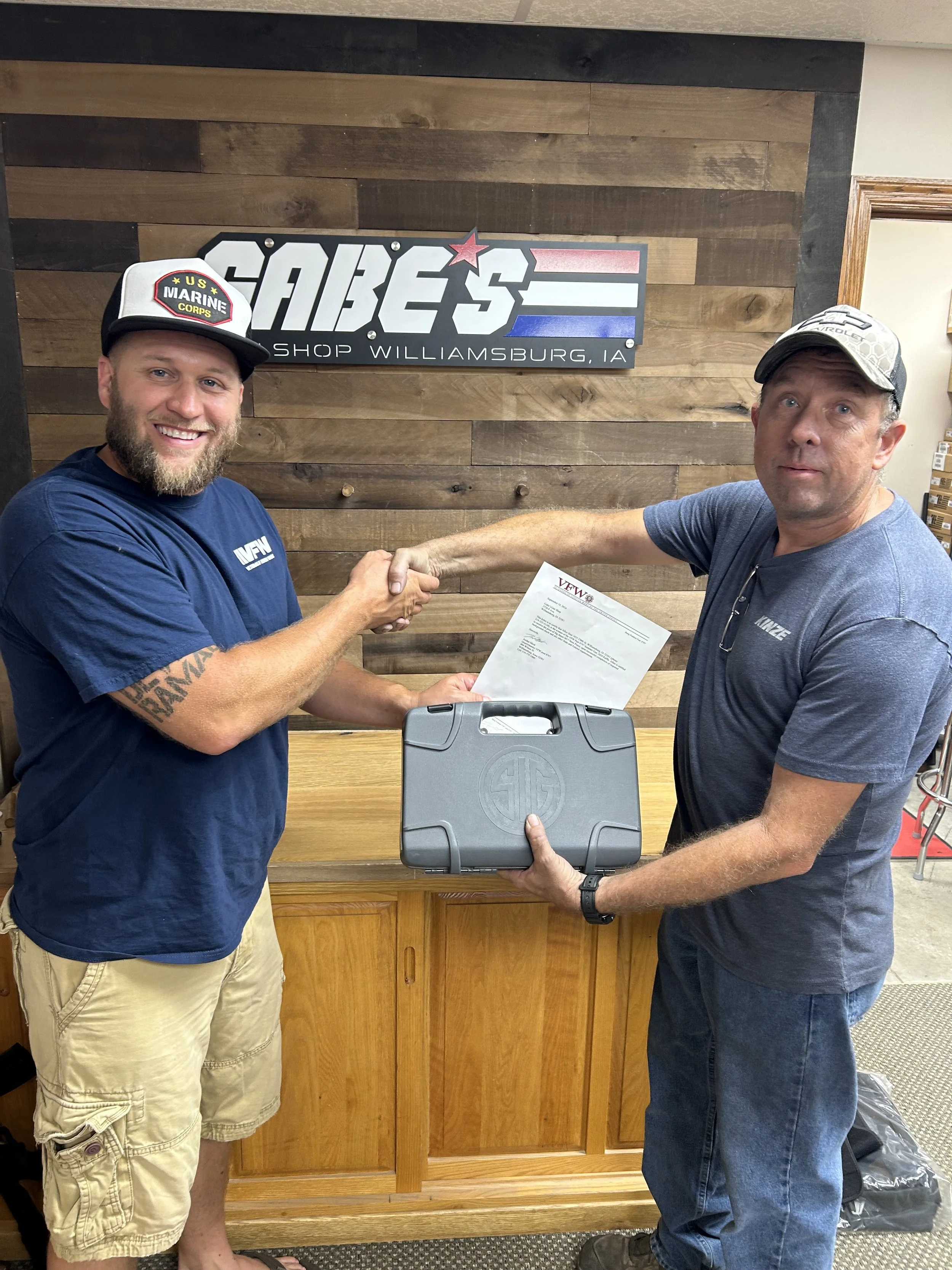 Two men shaking hands inside a store, holding a briefcase and an envelope, with a wooden wall and a sign that reads 'CABE'S SHOP WILLIAMSBURG, IA' in the background.