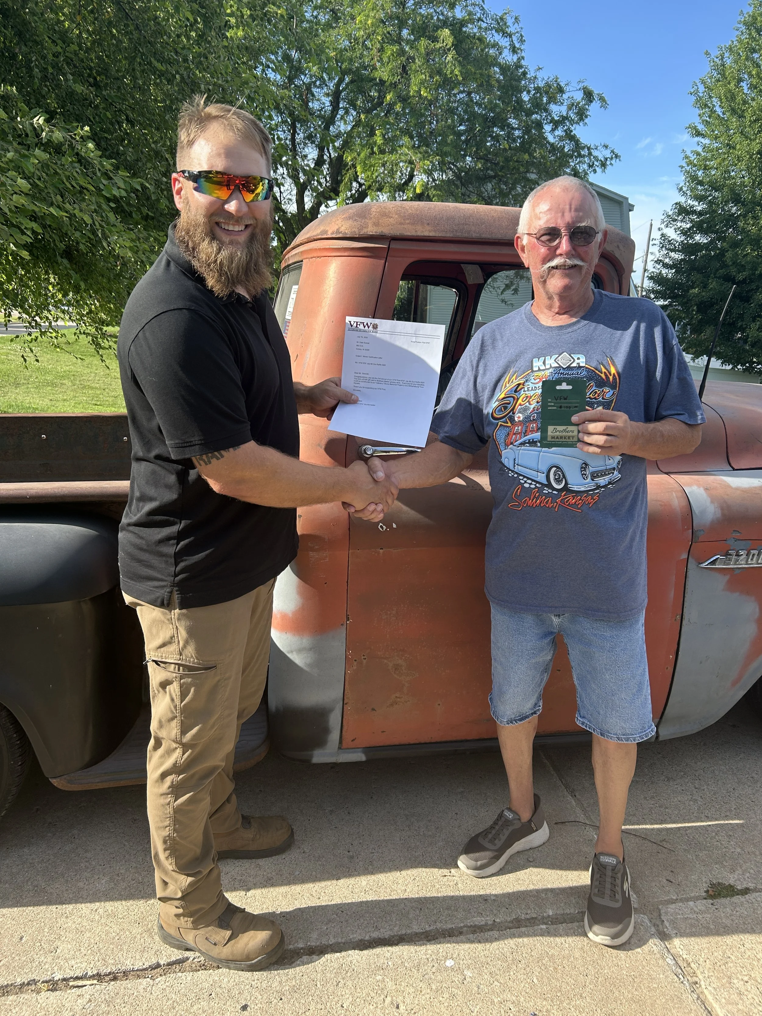 Two men shaking hands, one holding a certificate and the other holding a smartphone, standing in front of an old truck, outdoors with trees and blue sky.