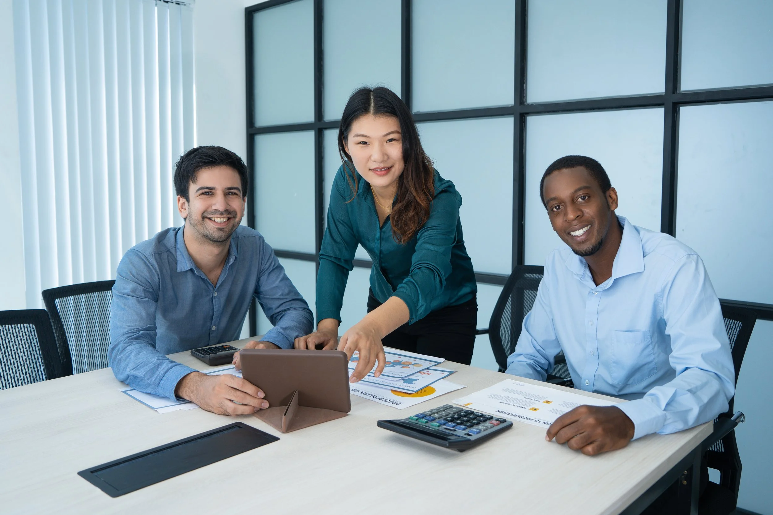 Three people in a business meeting, two men and one woman, interacting with documents and a tablet, in an office conference room.