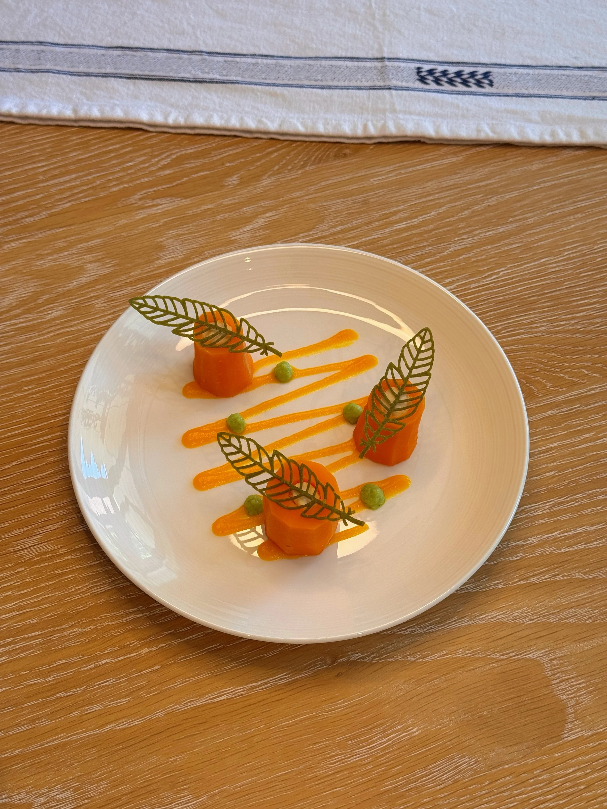 A white plate with three carrot cubes topped with decorative green leaves, green pea-like spheres, and orange sauce, arranged in a triangular pattern on a wooden table.