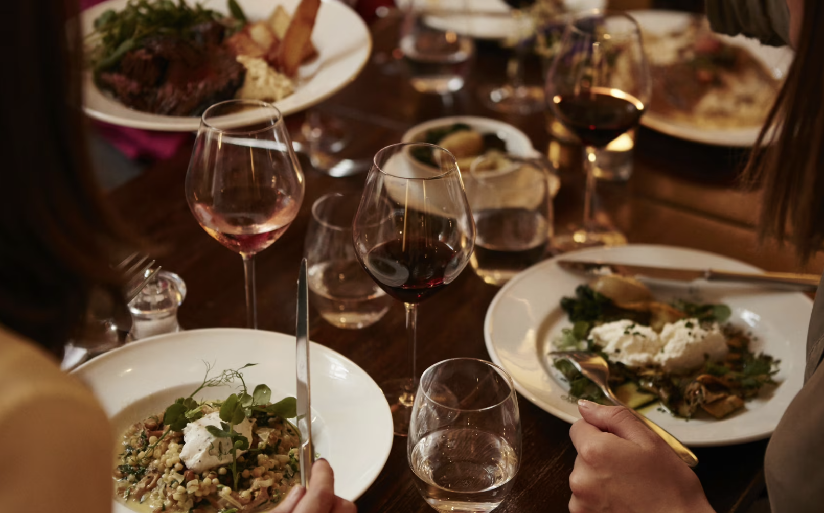 A dinner table with plates of food, several glasses of red and white wine, and people using utensils to eat.