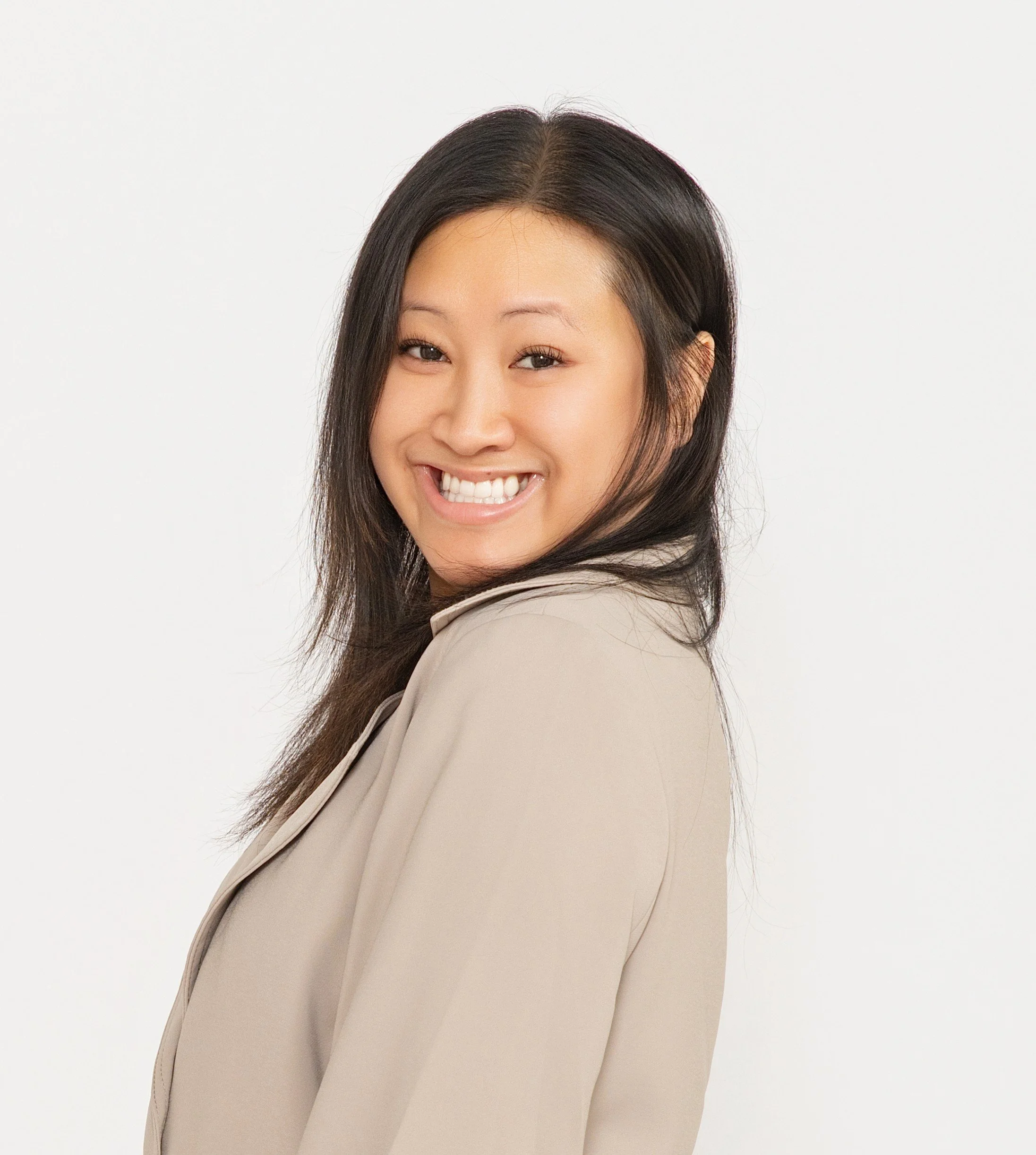 Close-up of a smiling woman with long dark hair, wearing a beige blazer, against a plain white background.
