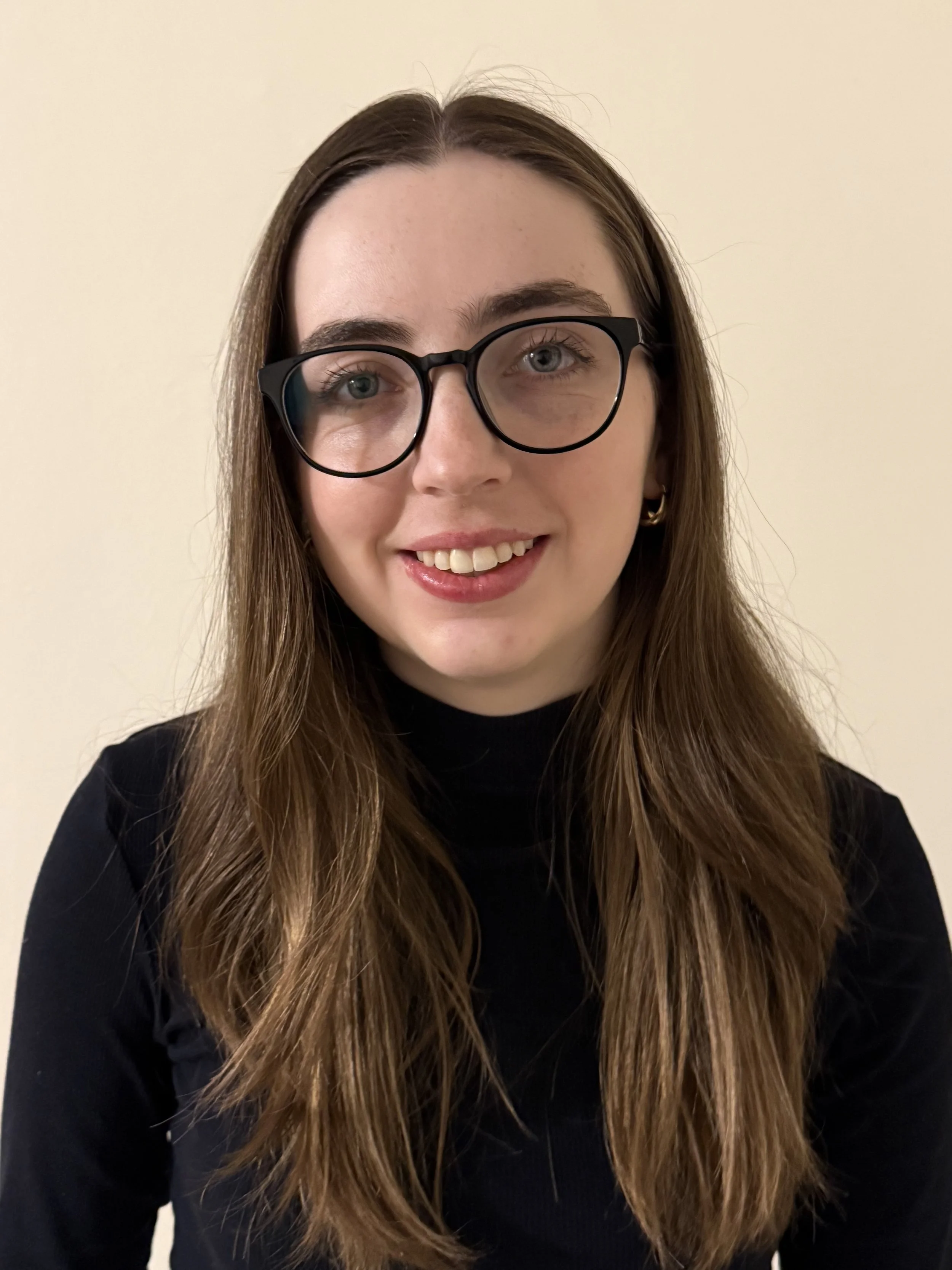 A young woman with long brown hair, wearing black glasses and a black top, smiling against a plain, light-colored wall.