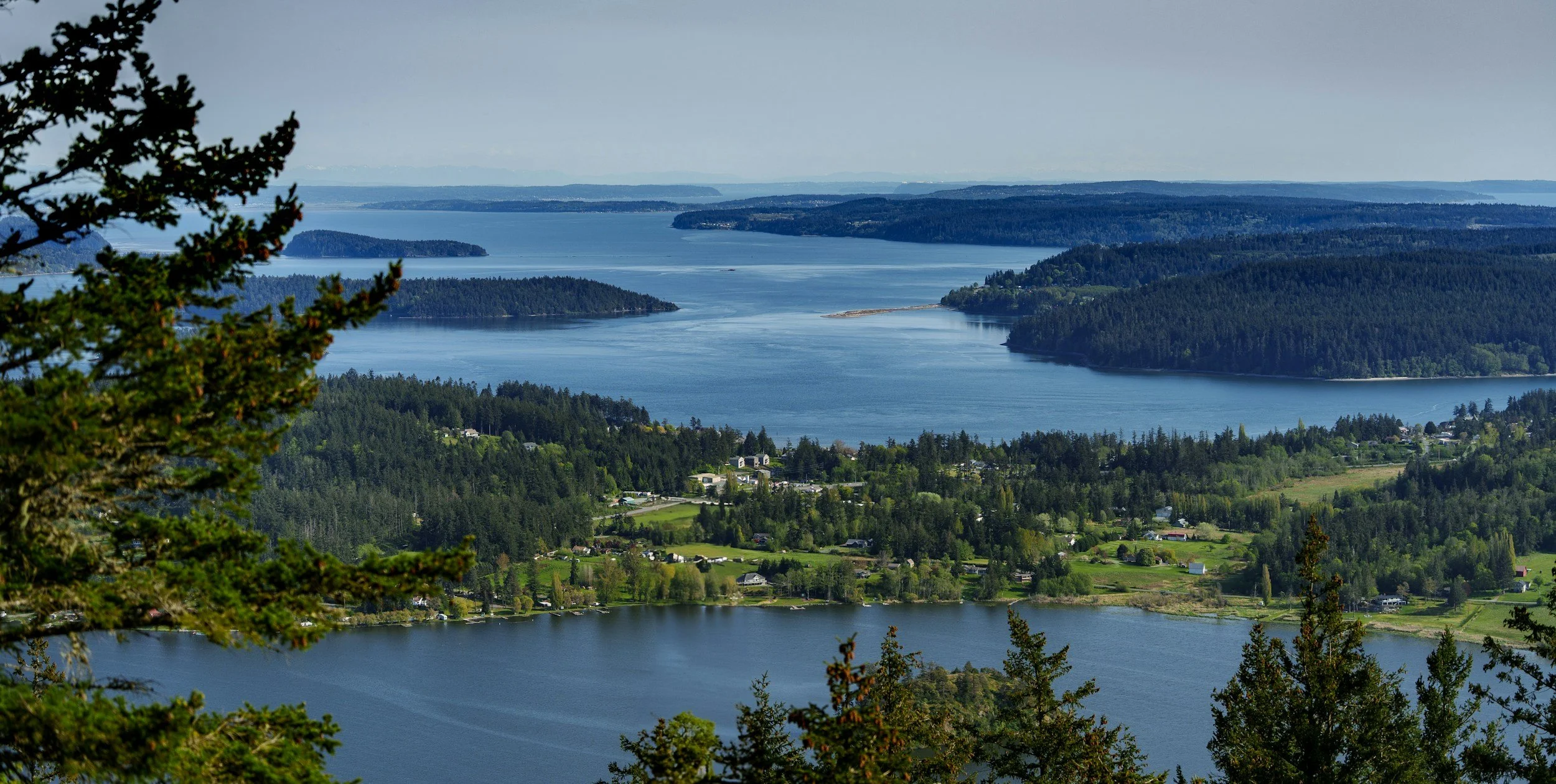 A panoramic view of a large body of water with several islands and peninsulas, surrounded by forests and a small residential area with houses, trees, and green fields in the foreground.