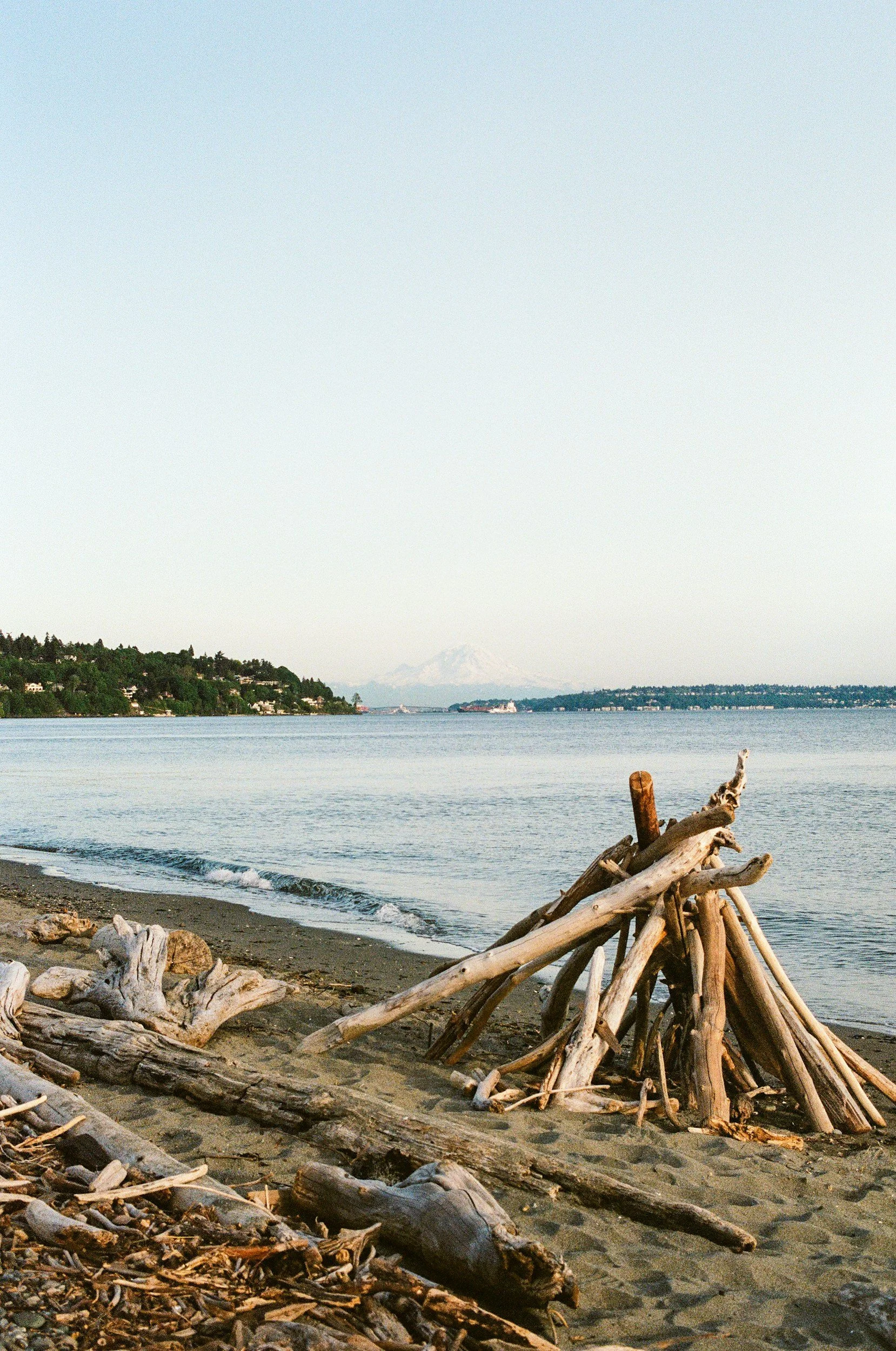 A beach with driftwood arranged in a teepee shape and smaller pieces scattered on the sand. Calm water extends to the horizon, with a landmass on the left and a faint snow-capped mountain in the distance.