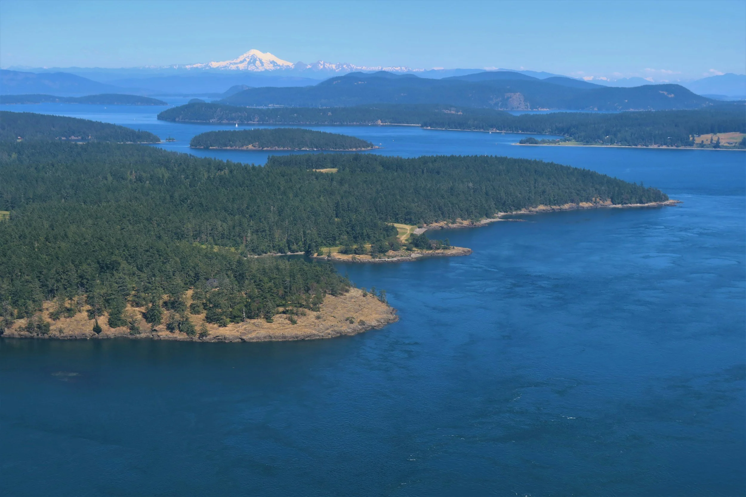 Aerial view of a lake with forested islands and surrounding mountains, including a snow-capped peak in the distance under a clear blue sky.