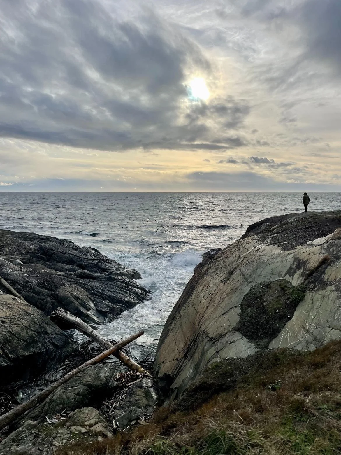 A person standing on a rocky coastline, facing the ocean with cloudy skies overhead and the sun partially obscured by clouds.