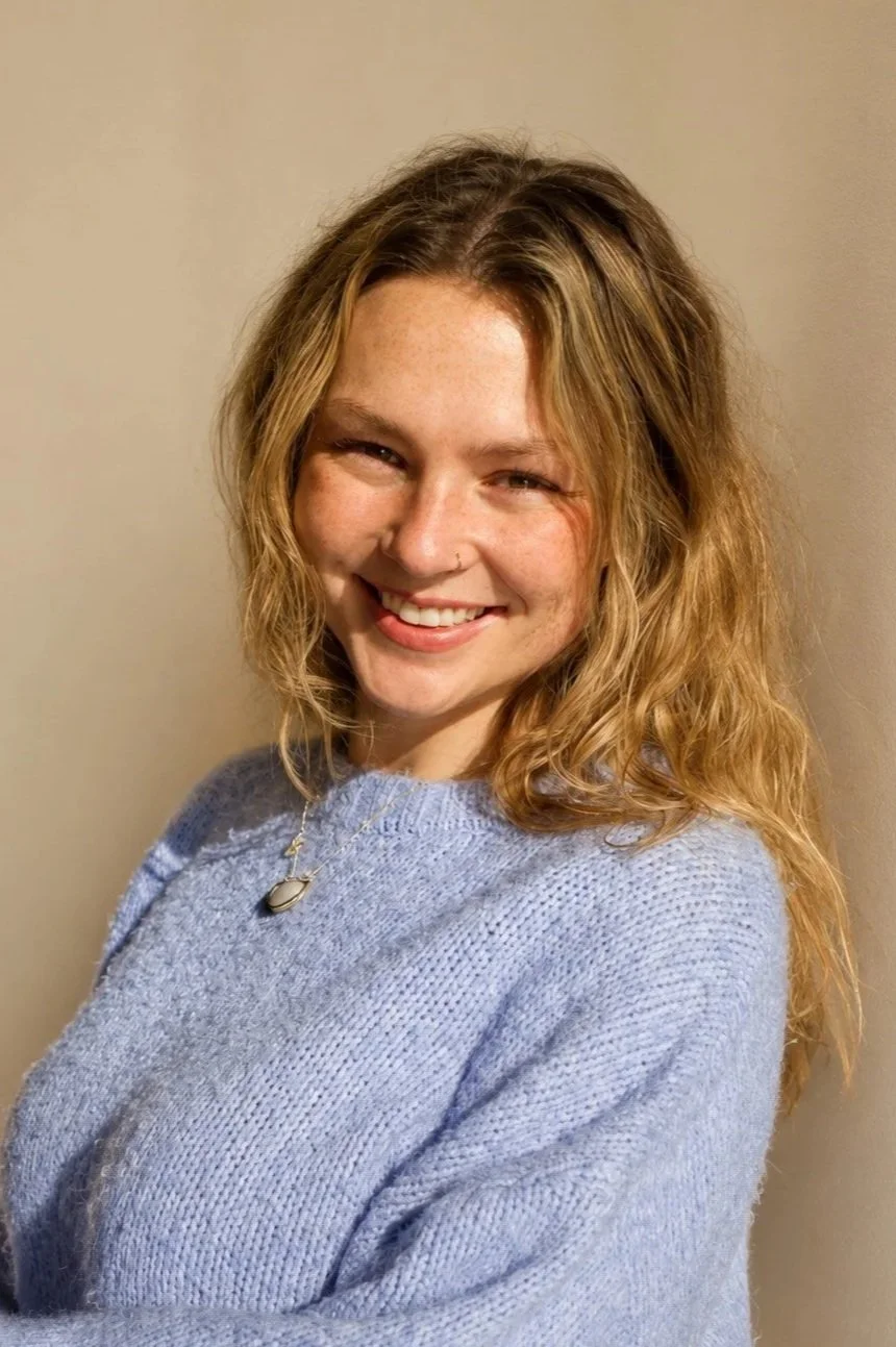 A young woman with wavy, shoulder-length blonde hair, smiling and wearing a light blue sweater and a necklace, standing against a plain beige wall.