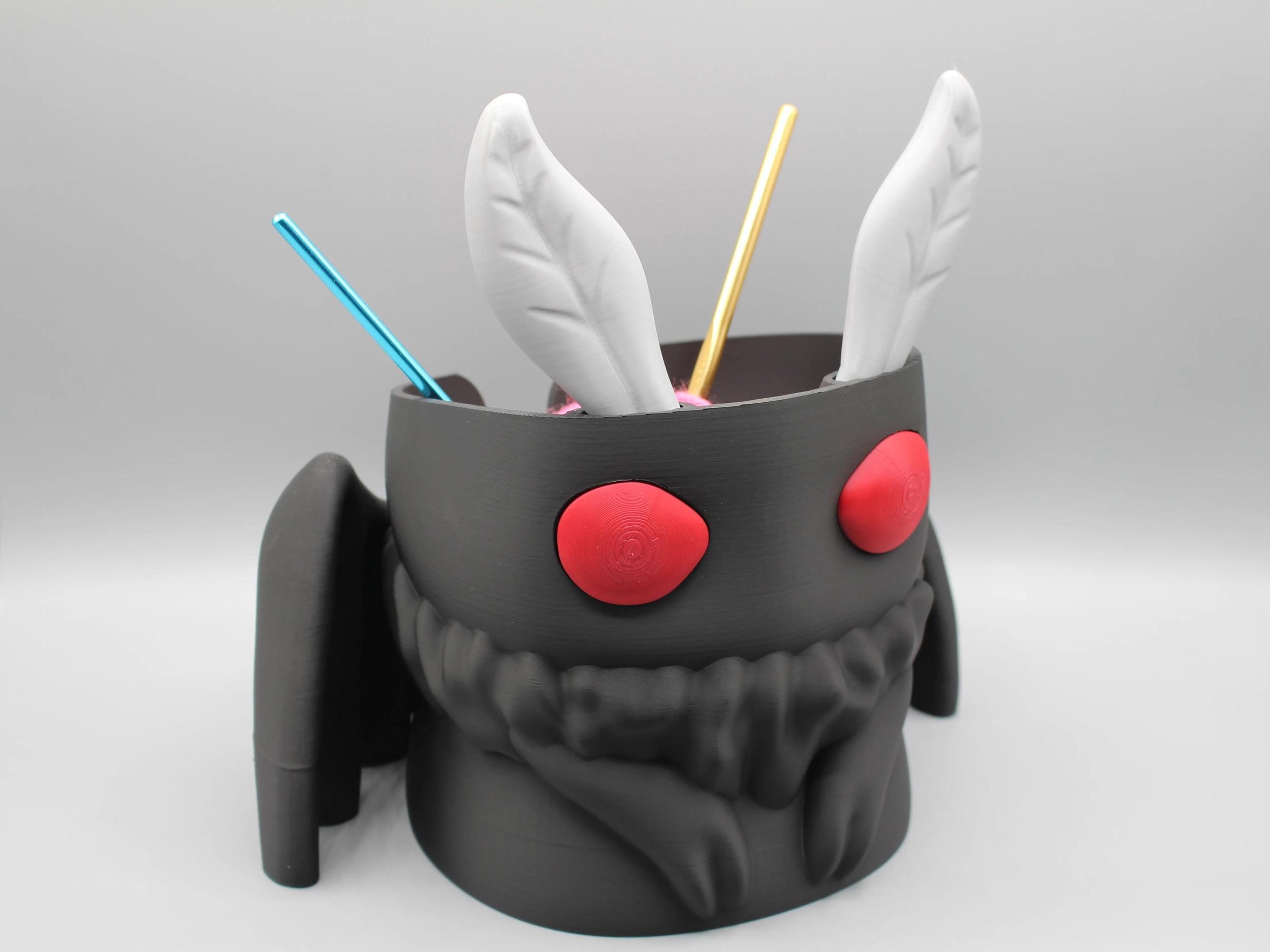 A 3D-printed black and gray mug shaped like a cute robot with red buttons for eyes and black limbs, containing three white feathers and two colorful straws.