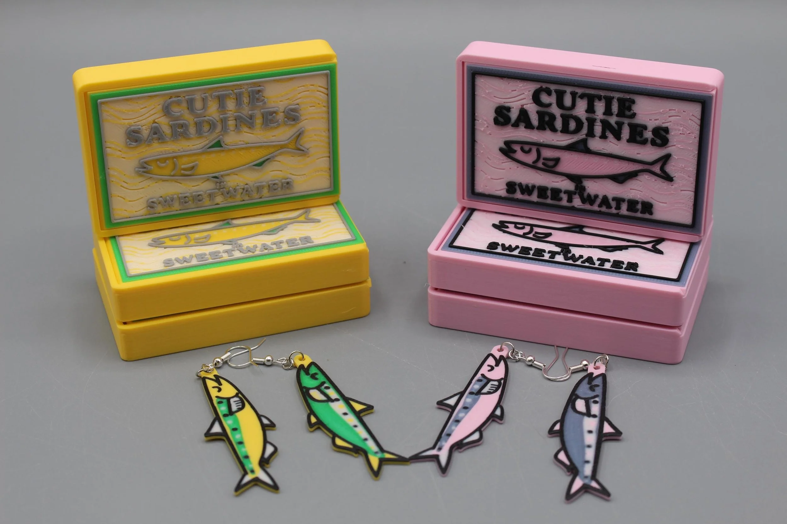 Colorful jewelry set with fish-shaped pendants and earrings in yellow, green, pink, and blue, displayed in yellow and pink boxes marked 'Cutie Sardines Sweetwater'.