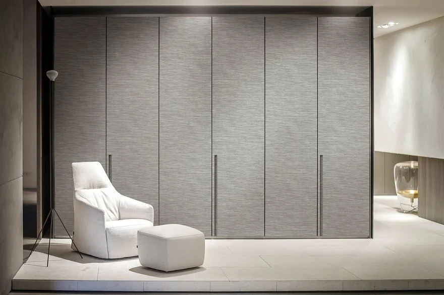 Modern minimalist room with a tall grey wardrobe, white armchair, matching ottoman, and a small side table with a glass vase. Soft lighting enhances the neutral color palette.