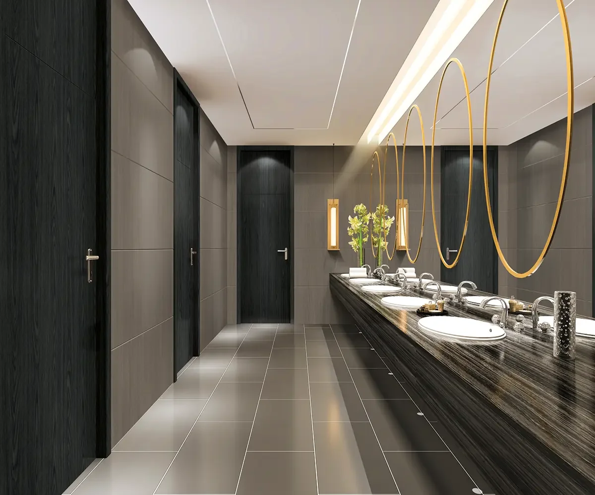 Modern men's or women's restroom with four sinks, large mirrors, and decorative gold circular frames on the wall. Gray tiled floor and walls, with black doors and a sleek dark wood countertop.