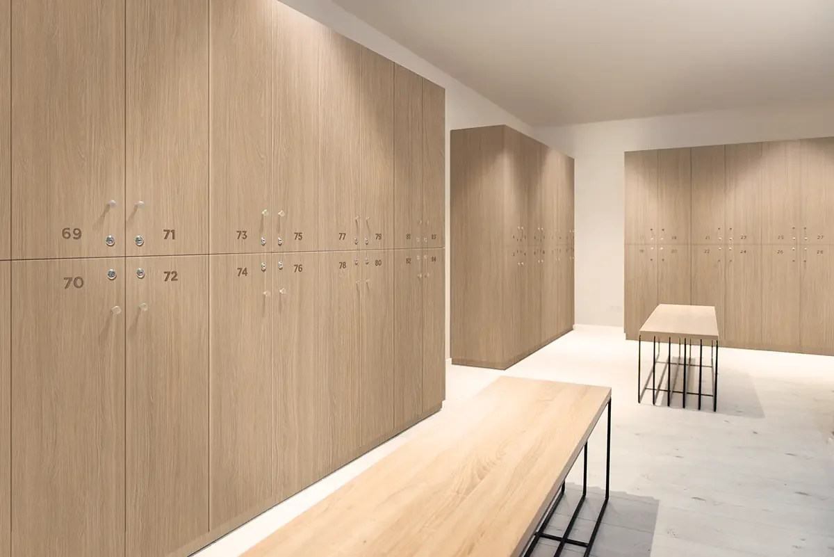 A locker room with light wooden lockers, some with small circular locks and numbered tags, a wooden bench, and a table with black metal legs.