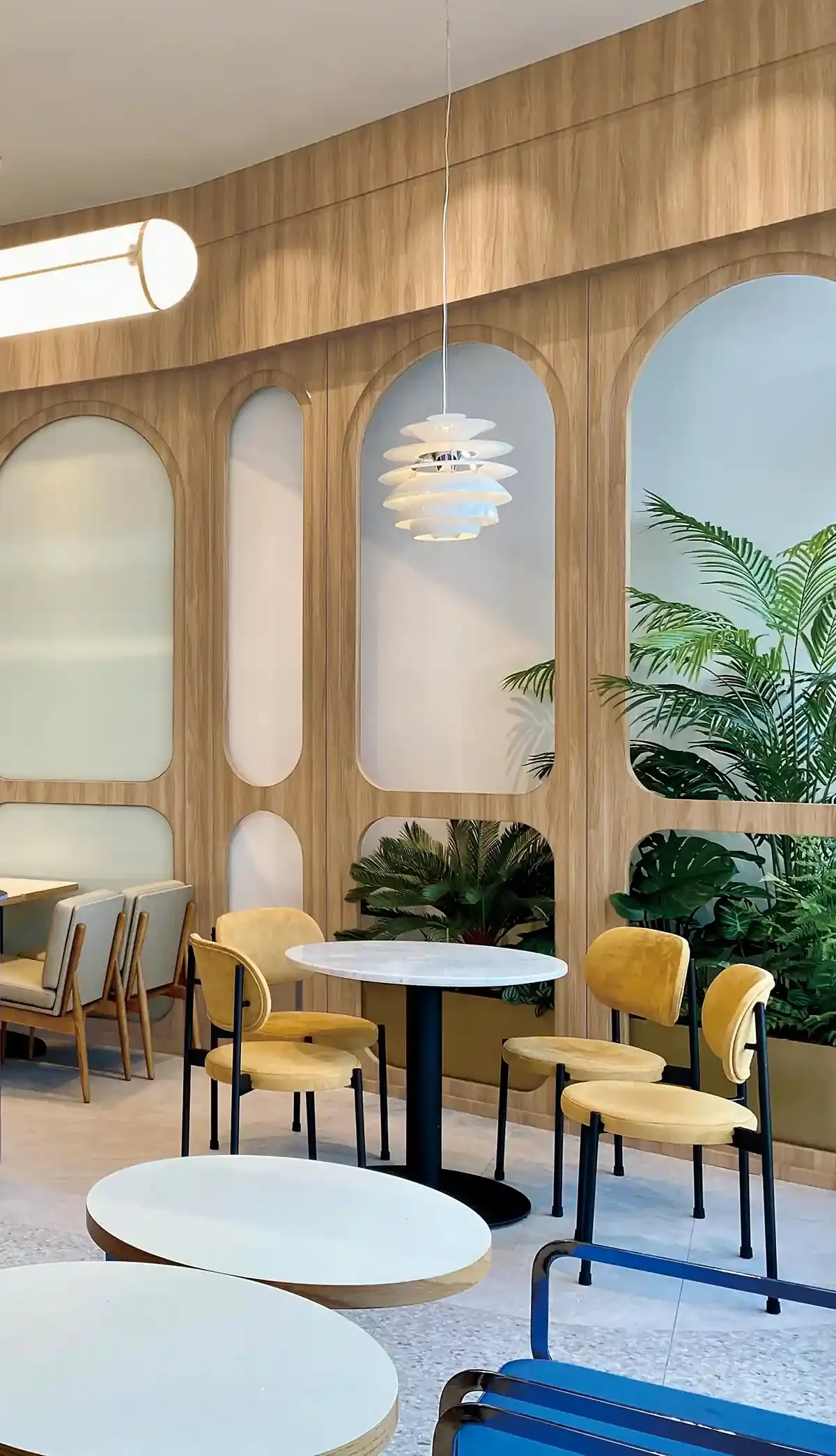 Interior of a modern café with wooden wall panels, yellow velvet chairs, white marble tables, and green indoor plants.