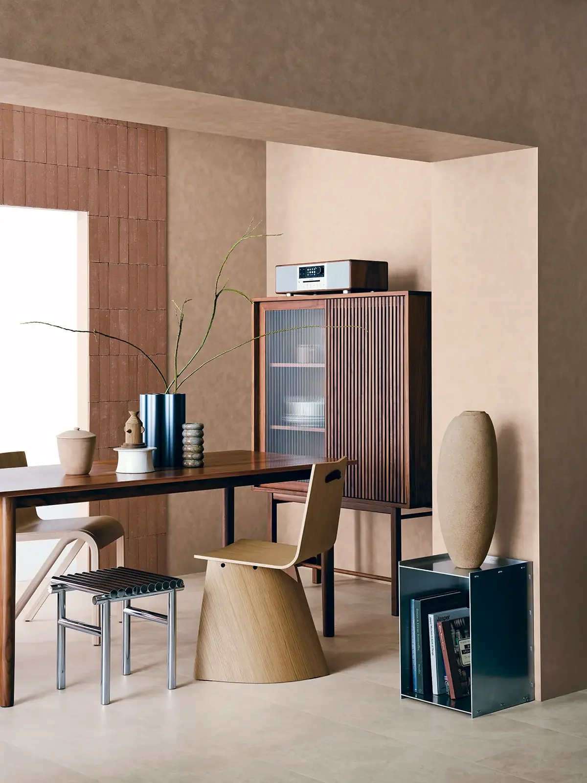Modern dining room with a wooden table, a light wood chair, a small black stool, large vases, and a cabinet with dishes inside, illuminated by natural light from a window.