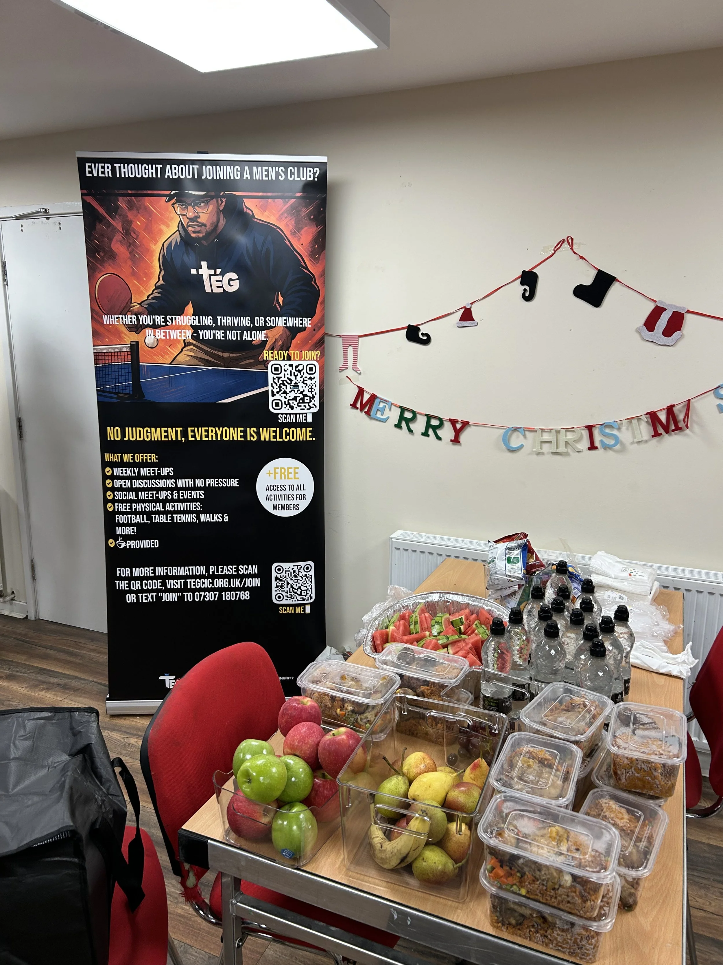 Table with apples, watermelon slices, bottled water, and food containers. In the background, a black and red banner for a men's club and a 'Merry Christmas' banner on the wall.