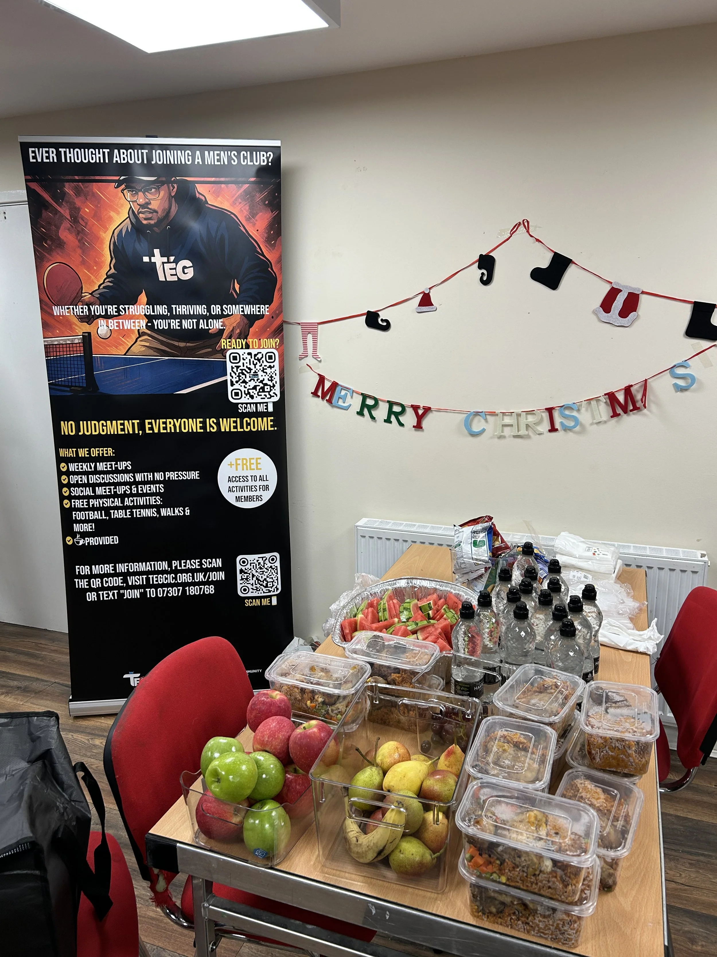 A room decorated for Christmas with a banner that reads 'MERRY CHRISTMAS' and a table filled with apples, water bottles, and prepared food containers. There is also a promotional poster for a men's club nearby.
