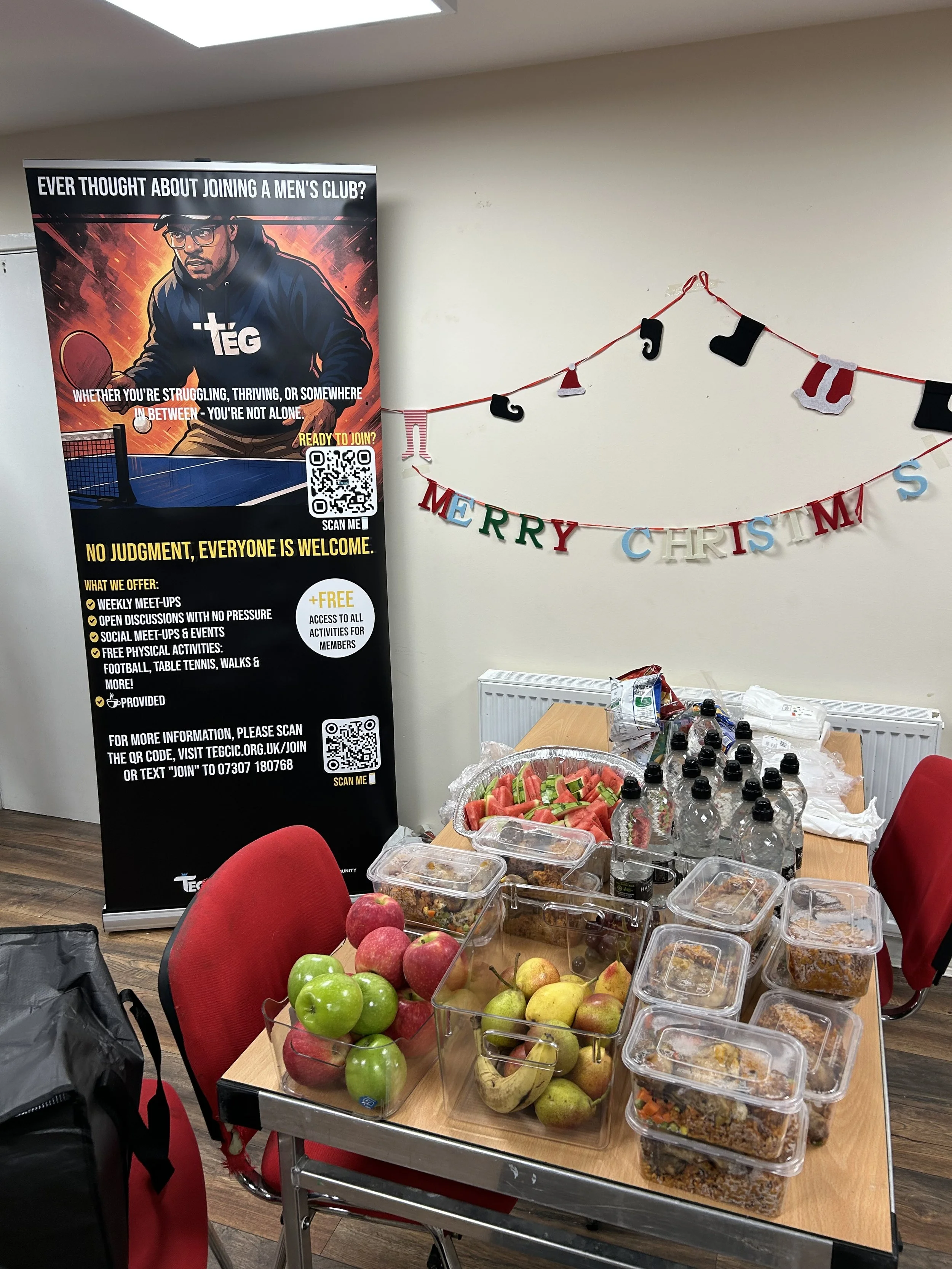 Table filled with various containers of food and bottles of water, with a holiday decoration on the wall and a promotional banner for a men's club in the background.