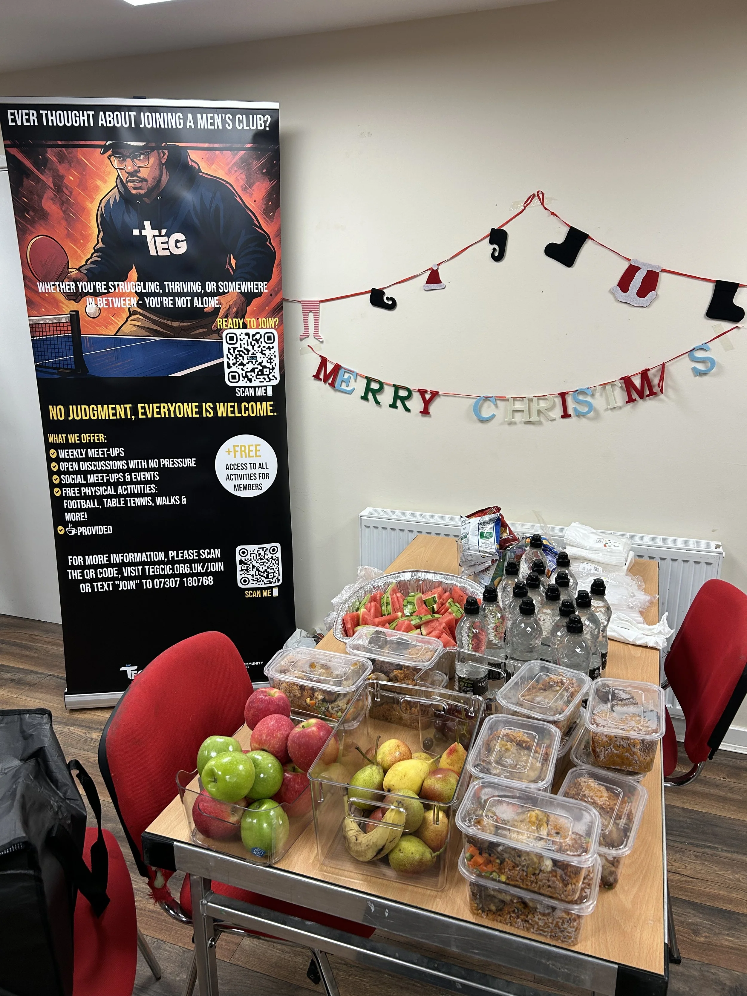Table with various snacks including water bottles, fruits, and pre-packaged foods, set up for a gathering in a room decorated with Christmas ornaments and a men's club recruitment banner.