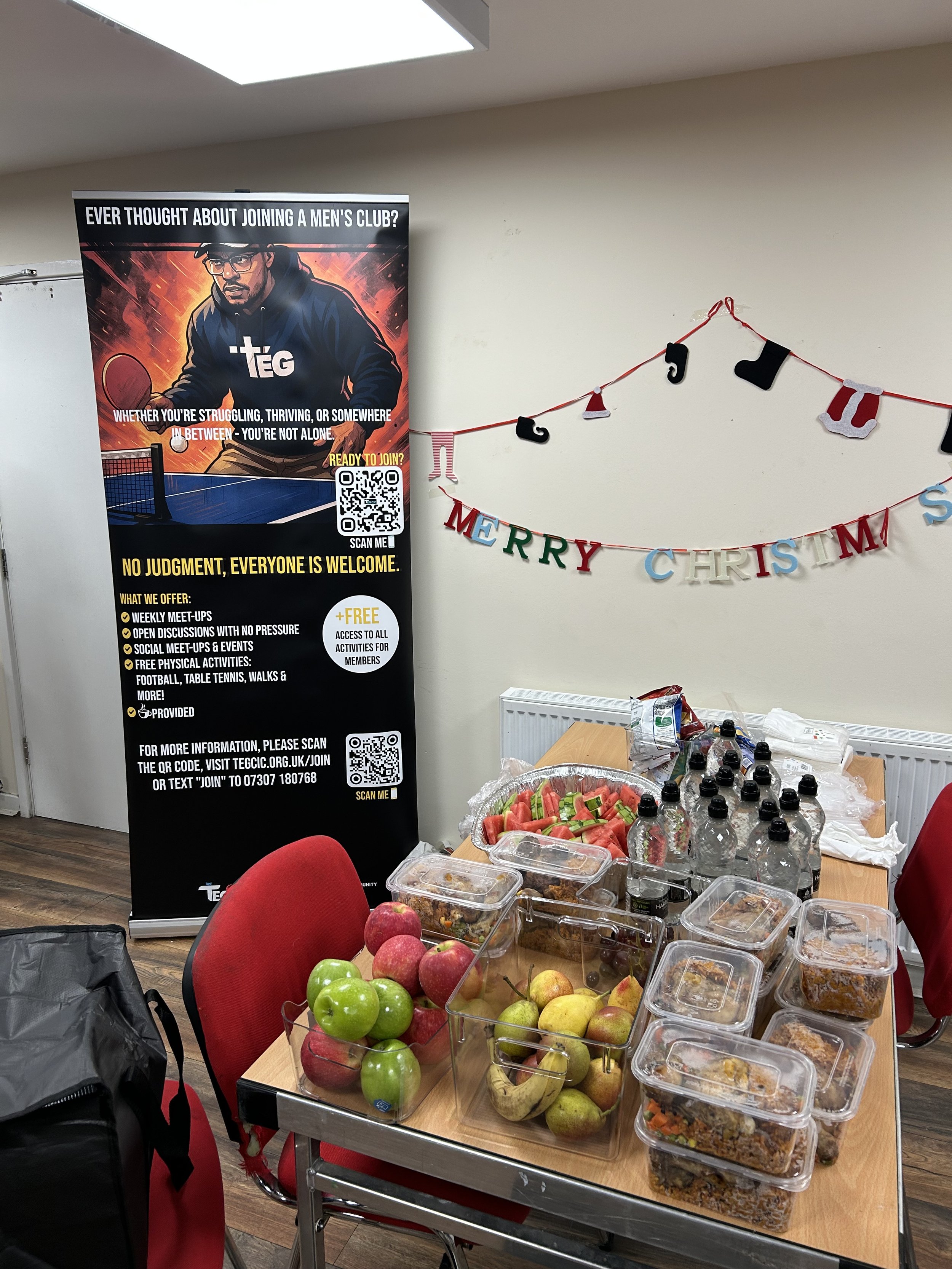 Table with assorted food containers, bottled water, and fresh fruit, set up for a gathering. Decor includes a 'Merry Christmas' banner on the wall, and a billboard promoting a men's club with a QR code.